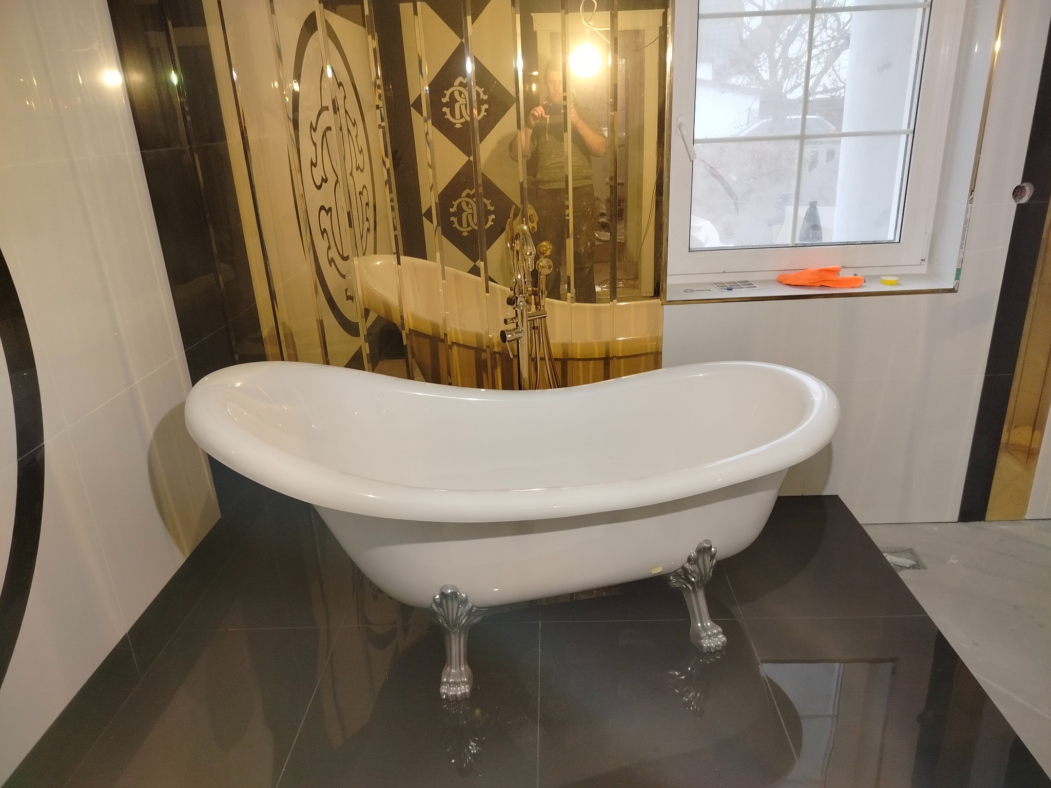 Elegancka, wolnostojąca wanna na lwich nóżkach w wykończonej łazience z lustrzanymi panelami i czarną podłogą z płytek. Widoczne akcesoria i odbicie fotografa.