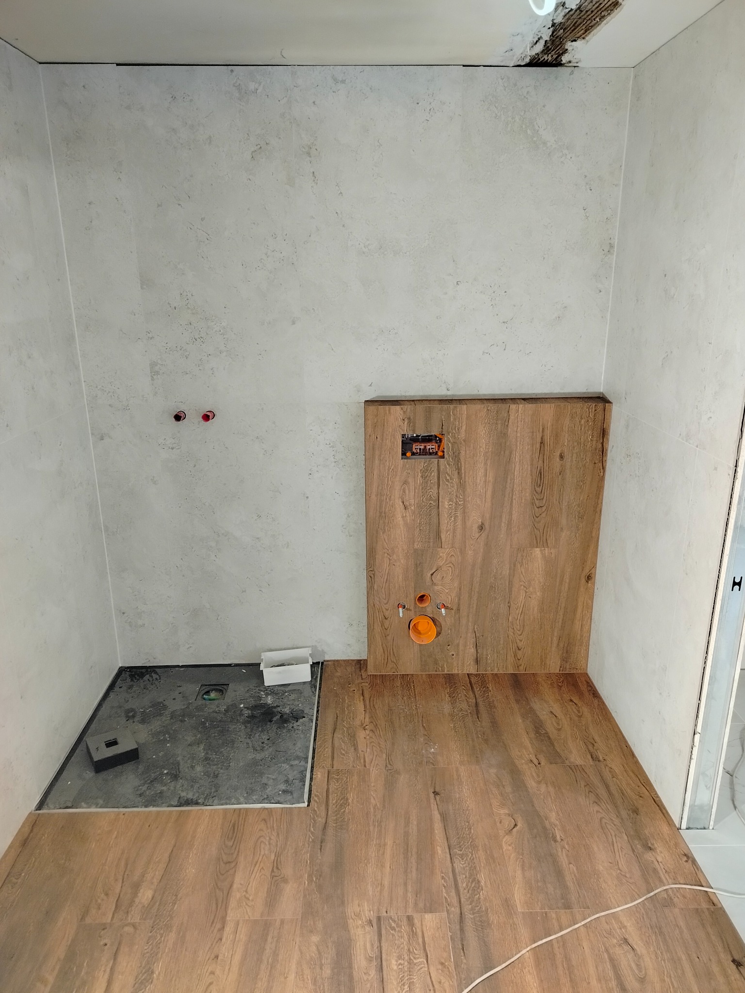Fragment łazienki w trakcie prac wykończeniowych: ułożone panele podłogowe imitujące drewno, ściany z szarych płytek i przygotowane miejsce na kabinę prysznicową.
