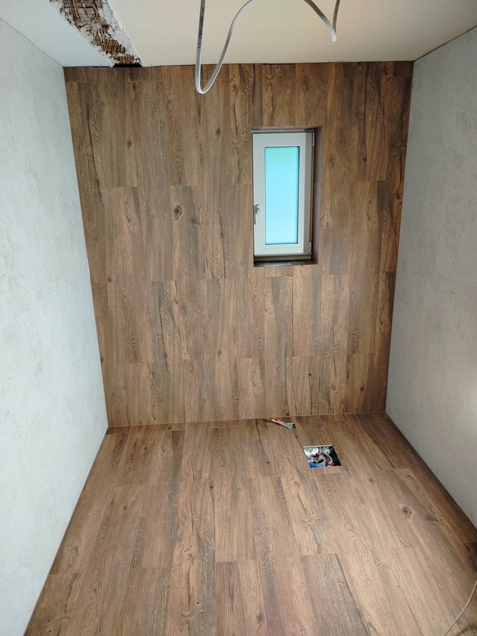 Wykończone wnętrze z panelami drewnopodobnymi na ścianach i podłodze, widoczne kable i puszka elektryczna, białe okno. Część sufitu wymaga wykończenia.