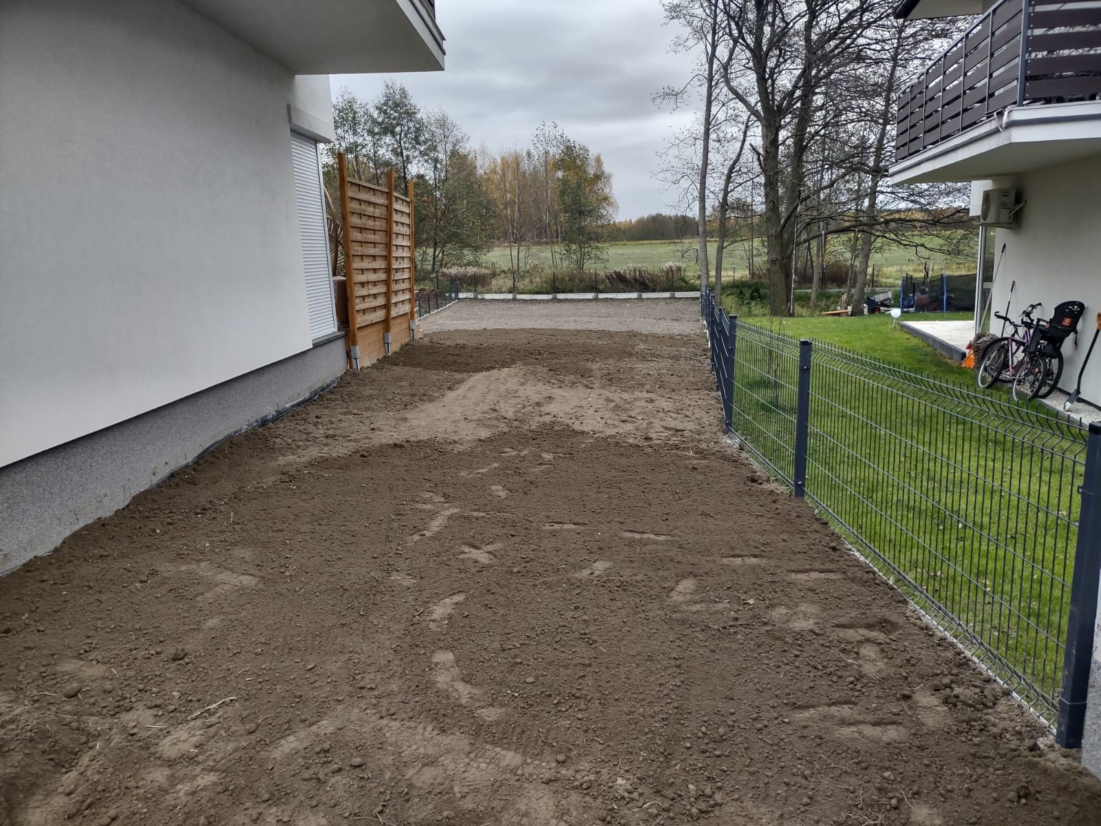 Przygotowany teren pod przyszły ogród, wyrównana ziemia, widoczne ogrodzenie panelowe, budynek z jasną elewacją i drewnianym elementem dekoracyjnym.