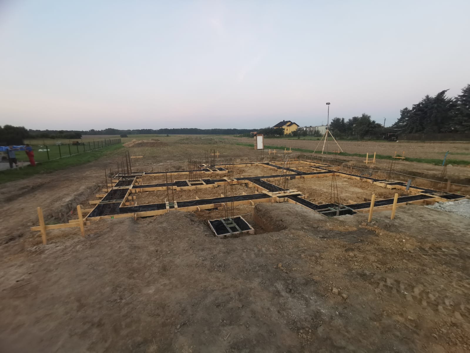 Zalane fundamenty domu jednorodzinnego z widocznym zbrojeniem, szalunkiem i izolacją, na tle pola i zabudowań, wczesnym rankiem.