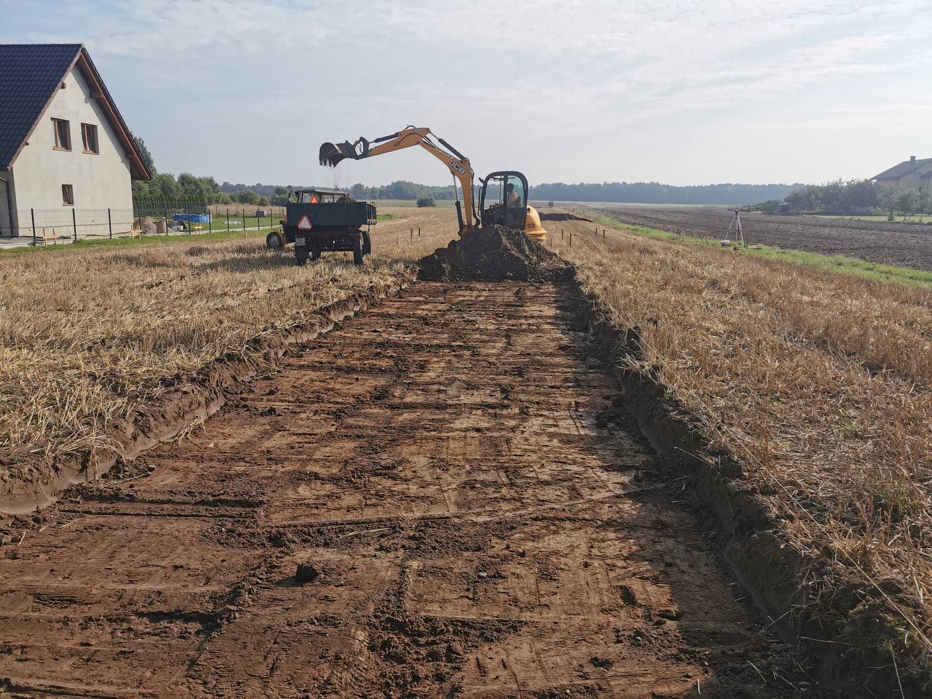 Żółta koparka JCB w trakcie wykopu rowu na polu ze ścierniskiem, obok przyczepa rolnicza i dom w tle. Widoczne ślady opon i świeżo wykopana ziemia.