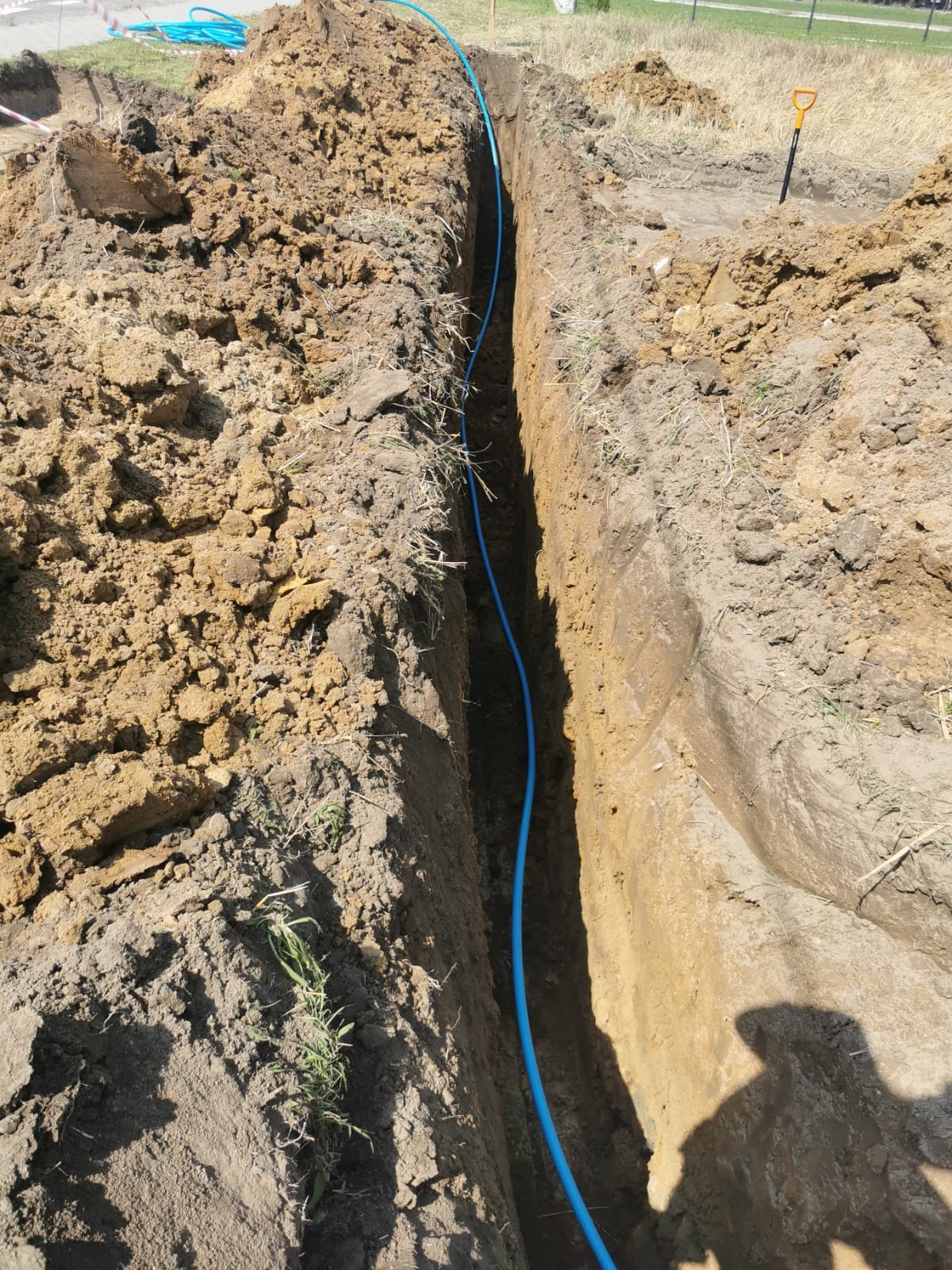 Wąski wykop w ziemi z niebieskim przewodem ułożonym wzdłuż niego. Ziemia po bokach wykopu, w tle trawa i częściowo widoczna łopata.