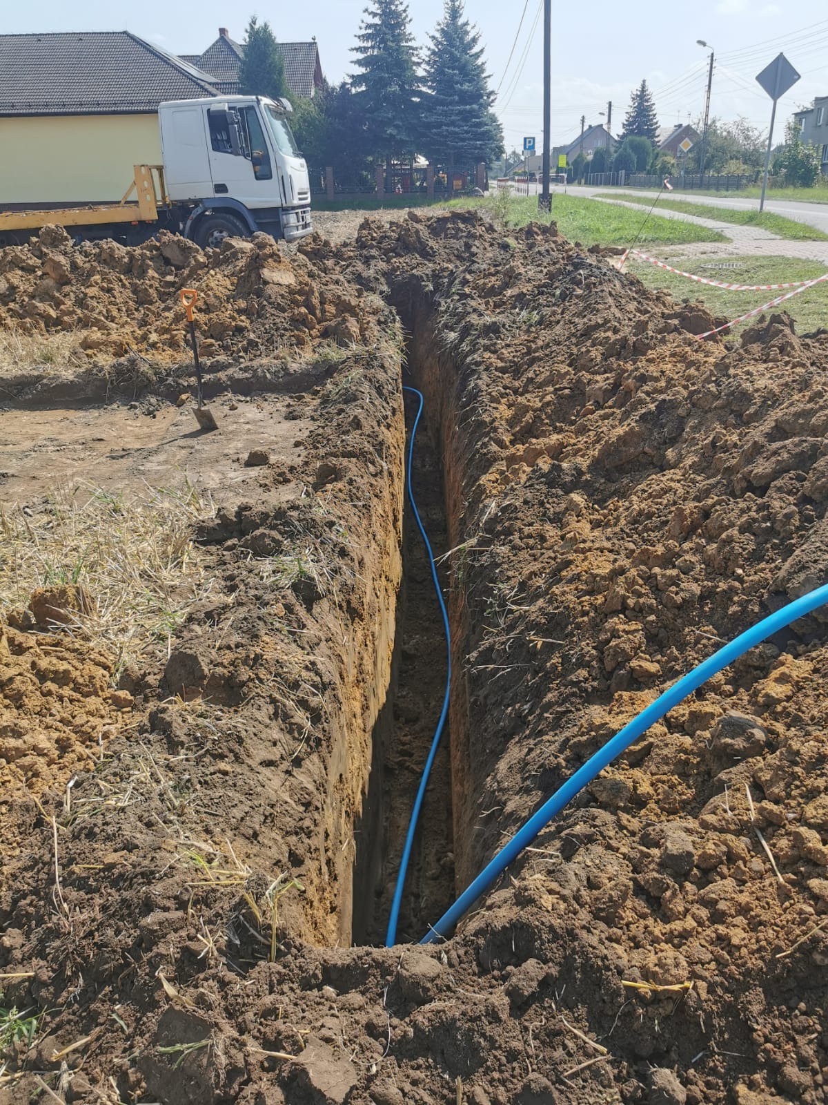 Wąski wykop w ziemi z niebieską rurą w środku, na tle krajobrazu wiejskiego z białym samochodem ciężarowym i domami. Wykop pod instalacje.