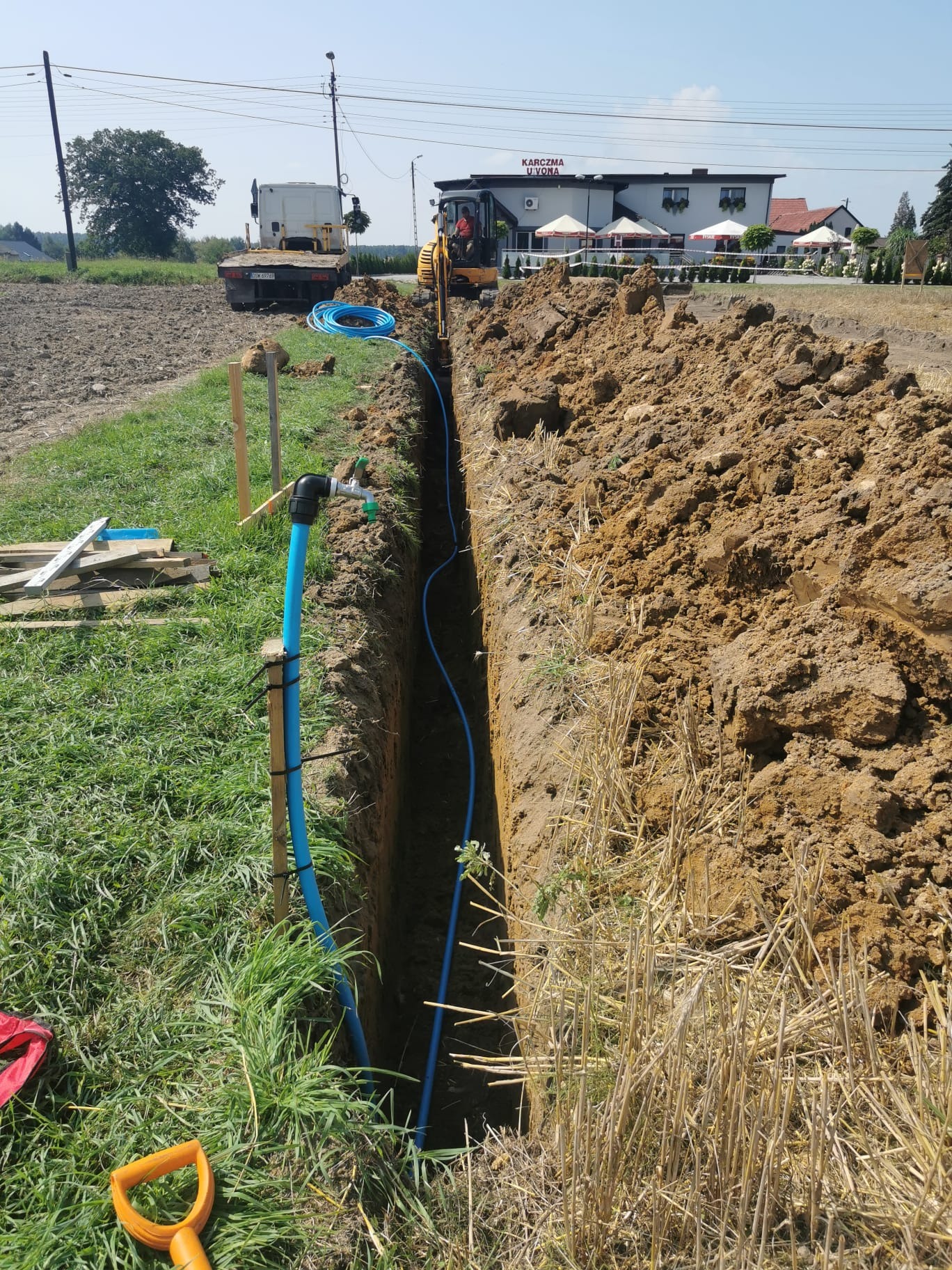 Długi, wąski wykop w ziemi z niebieską rurą instalacyjną w środku. W tle widoczny mały traktor, ciężarówka i budynek.