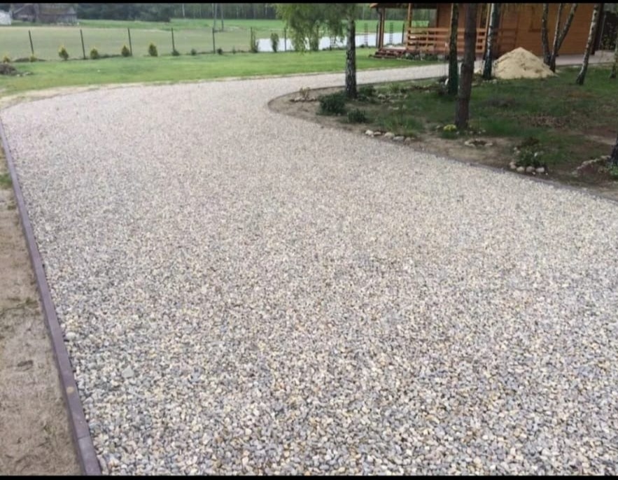 Utwardzona droga z jasnego żwiru, obrzeża z kostki. Widoczny fragment trawnika i drzewa w tle. Teren prywatny z elementami architektury ogrodowej.