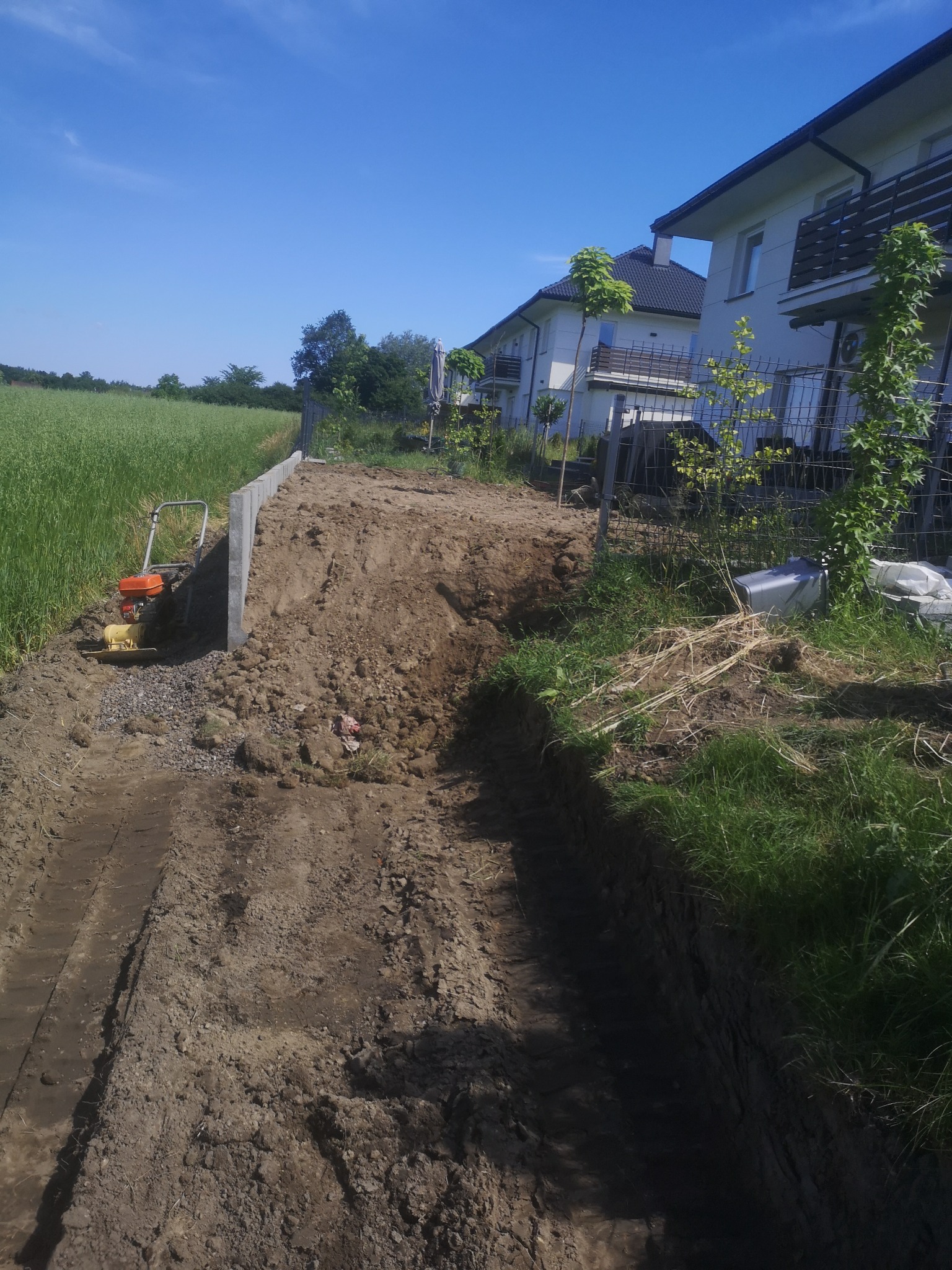Wykop pod fundament z widoczną ubijarką mechaniczną, sąsiadujący z zielonym polem i szeregiem domów w tle. Prace ziemne w toku, widoczne ślady opon.