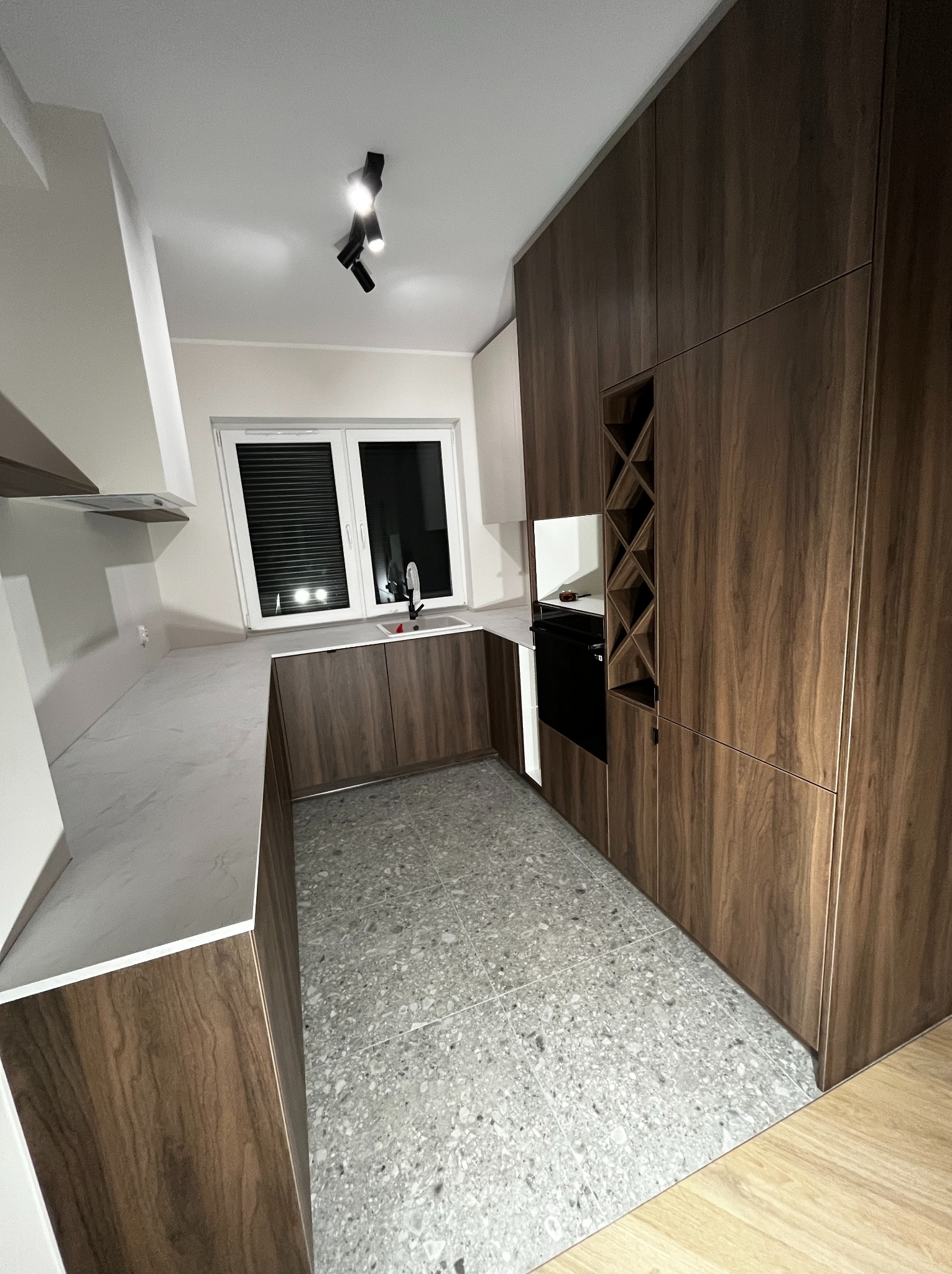 Nowoczesna kuchnia z drewnianymi frontami, szarym blatem i podłogą, oświetlona punktowymi lampami. W zabudowie regał na wino i piekarnik. Minimalistyczny design.