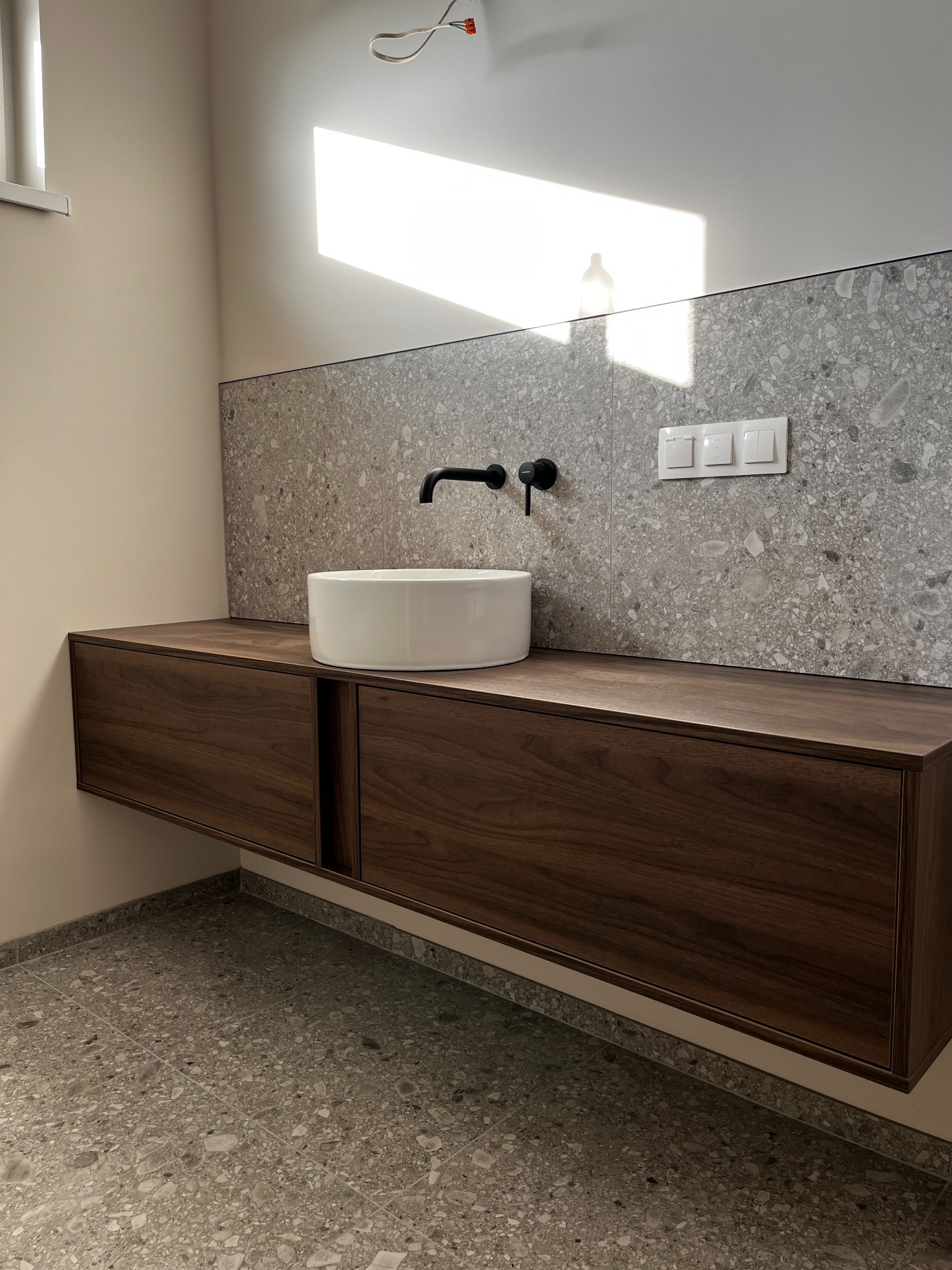 Nowoczesna szafka łazienkowa z drewna orzechowego, z okrągłą umywalką nablatową, czarną armaturą i szarą mozaiką. Promienie słoneczne podkreślają minimalistyczny design.