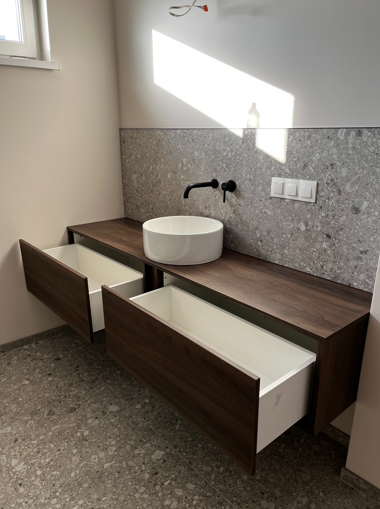 Nowoczesna łazienka z blatem z ciemnego drewna, okrągłą białą umywalką i otwartymi szufladami. Ściana wyłożona płytkami imitującymi lastryko. Widoczne czarne bateria i włączniki.