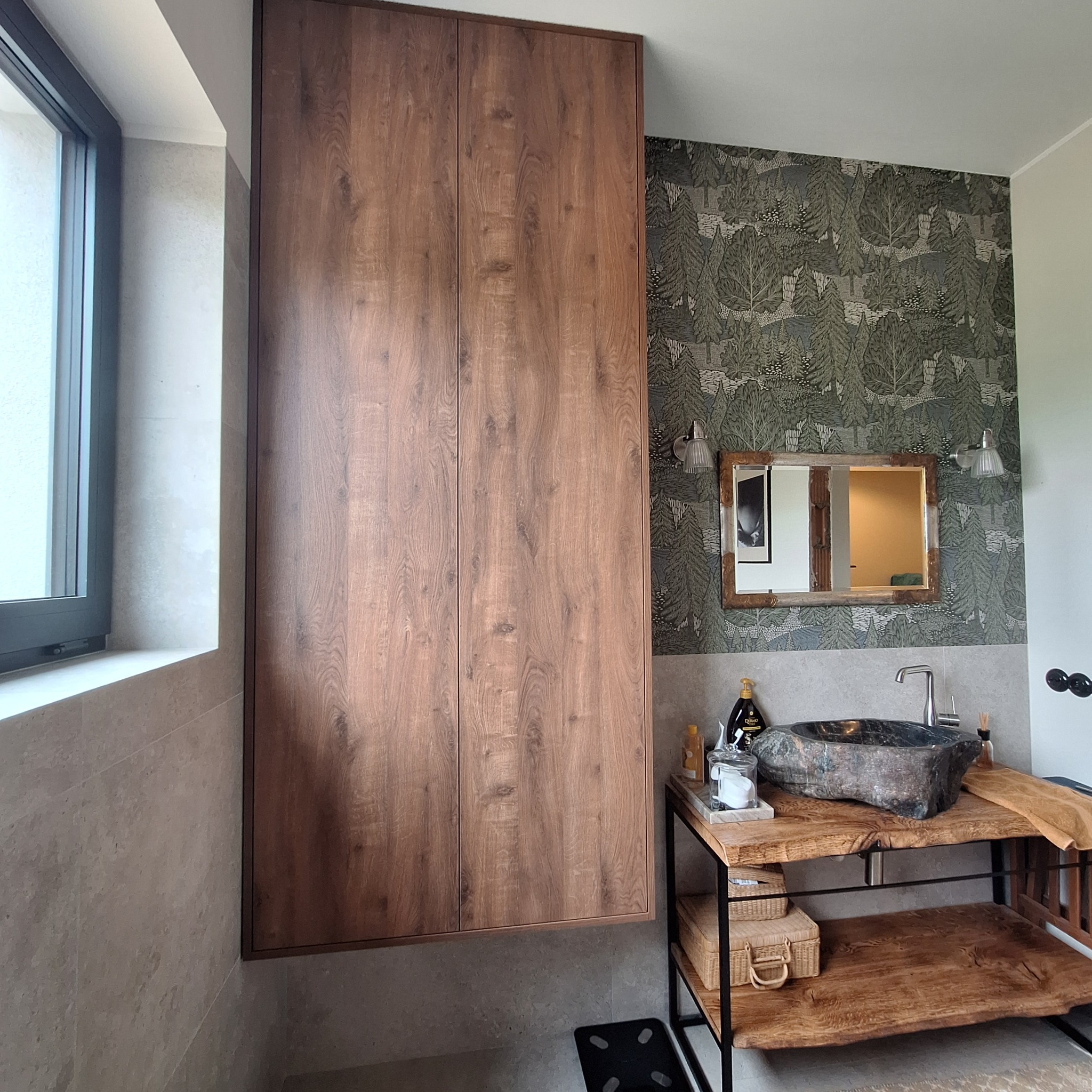 Wysoka, drewniana szafa wnękowa z dwiema pionowymi liniami podziału, w łazience z kamienną umywalką i wzorzystą tapetą w zielono-szare drzewa.