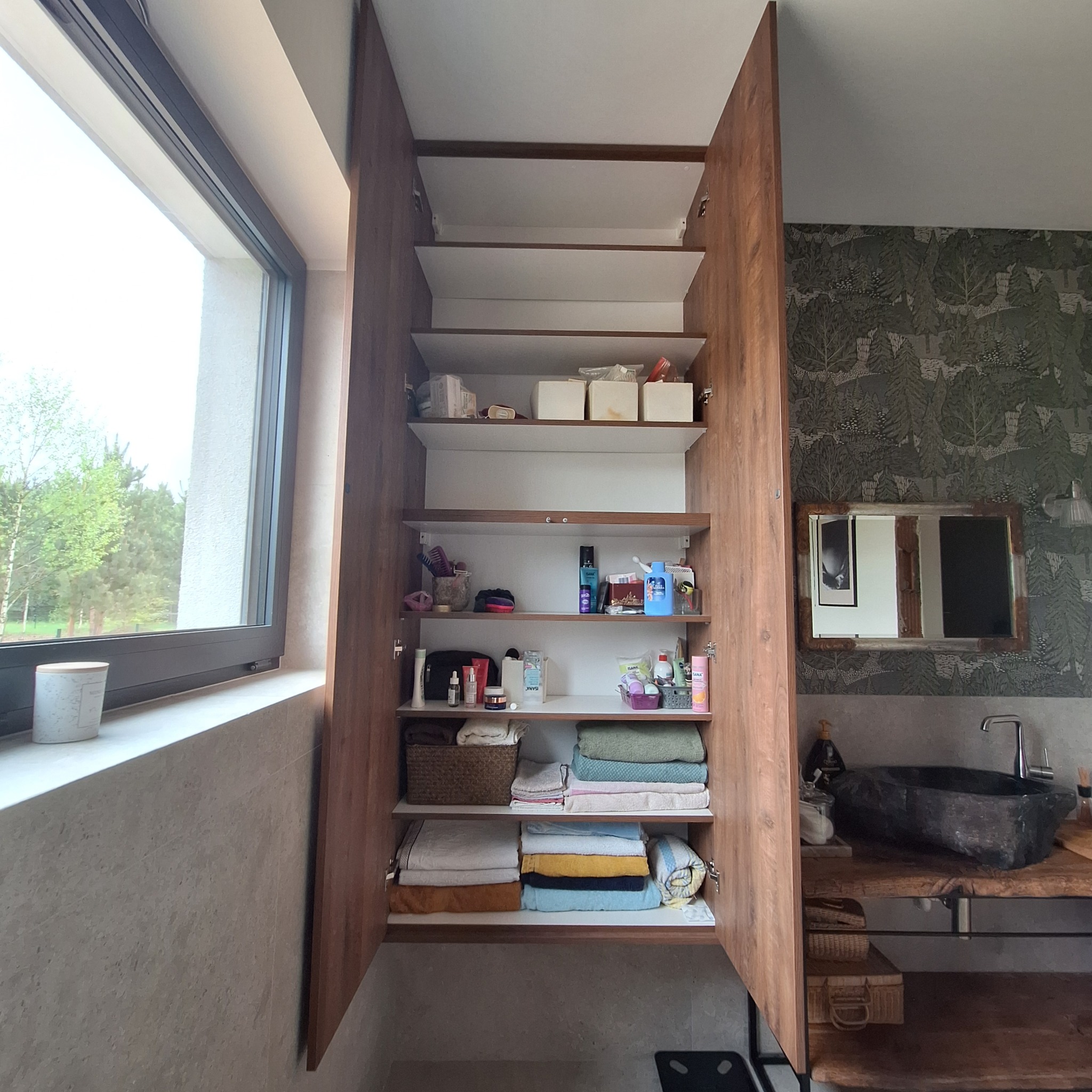 Otwarta szafa wnękowa z półkami wypełnionymi kosmetykami i ręcznikami w łazience z umywalką z kamienia i wzorzystą tapetą w tle.