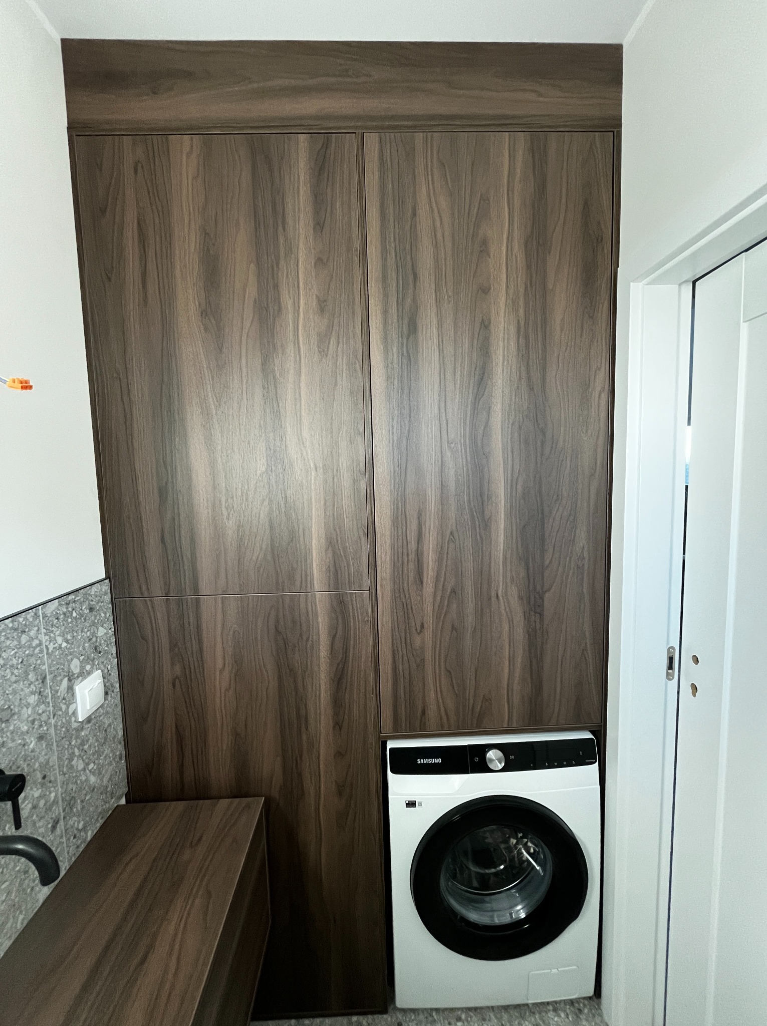 Wnękowa szafa z ciemnego drewna w łazience, z pralką Samsung u dołu. Minimalistyczny design, widoczny fragment umywalki i drzwi.