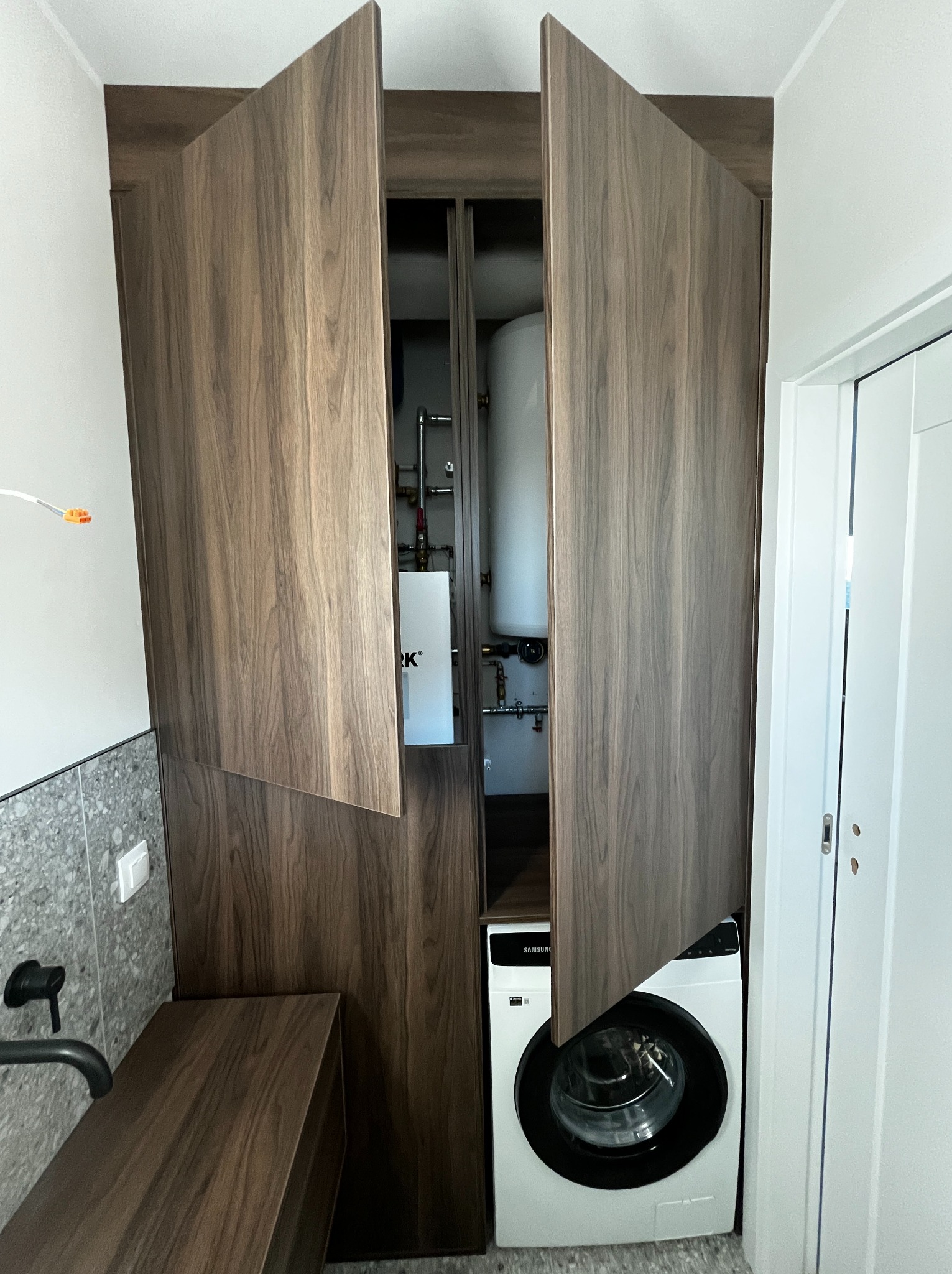 Szafa wnękowa z drewnianymi frontami, ukrywająca pralkę i bojler. Otwarta, ukazująca wnętrze. Minimalistyczny design, widoczna armatura.