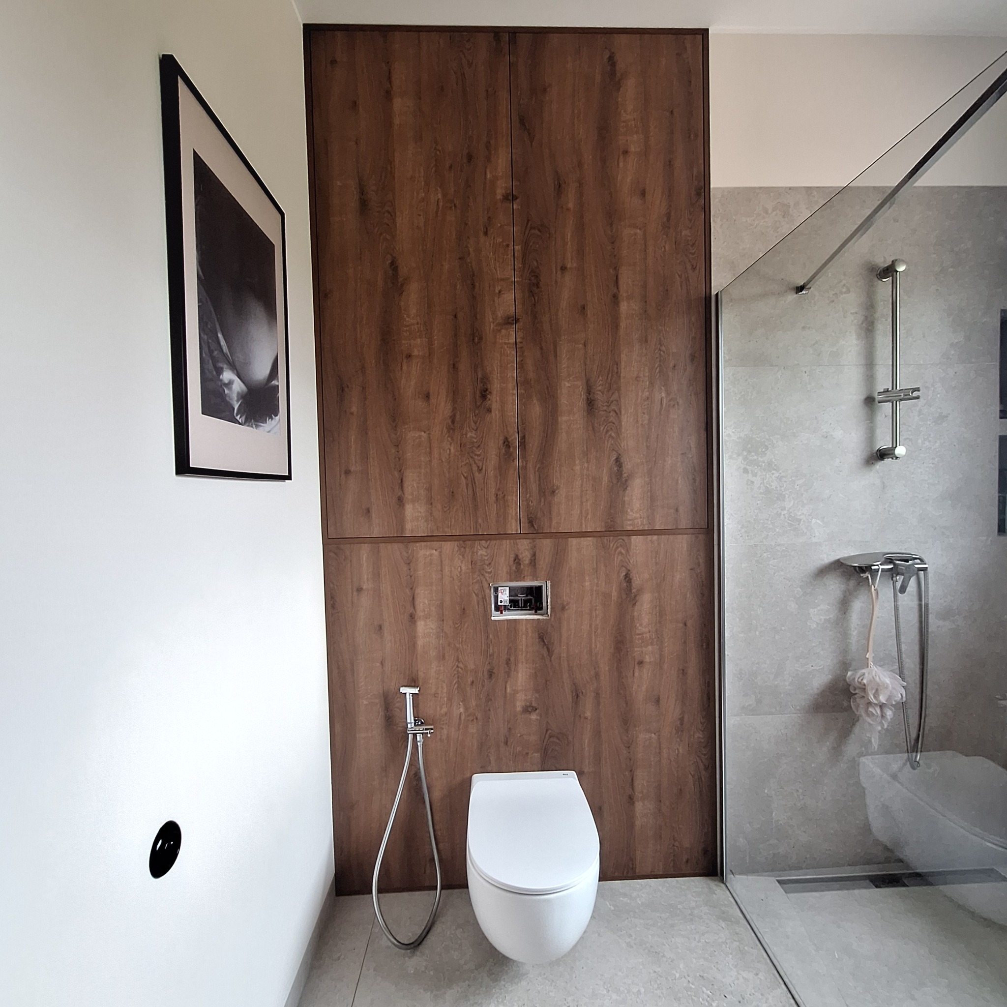 Nowoczesna łazienka z zabudową szaf w odcieniach drewna, wisząca toaleta z bidetką i kabina prysznicowa za szklaną ścianką. Minimalistyczny design.