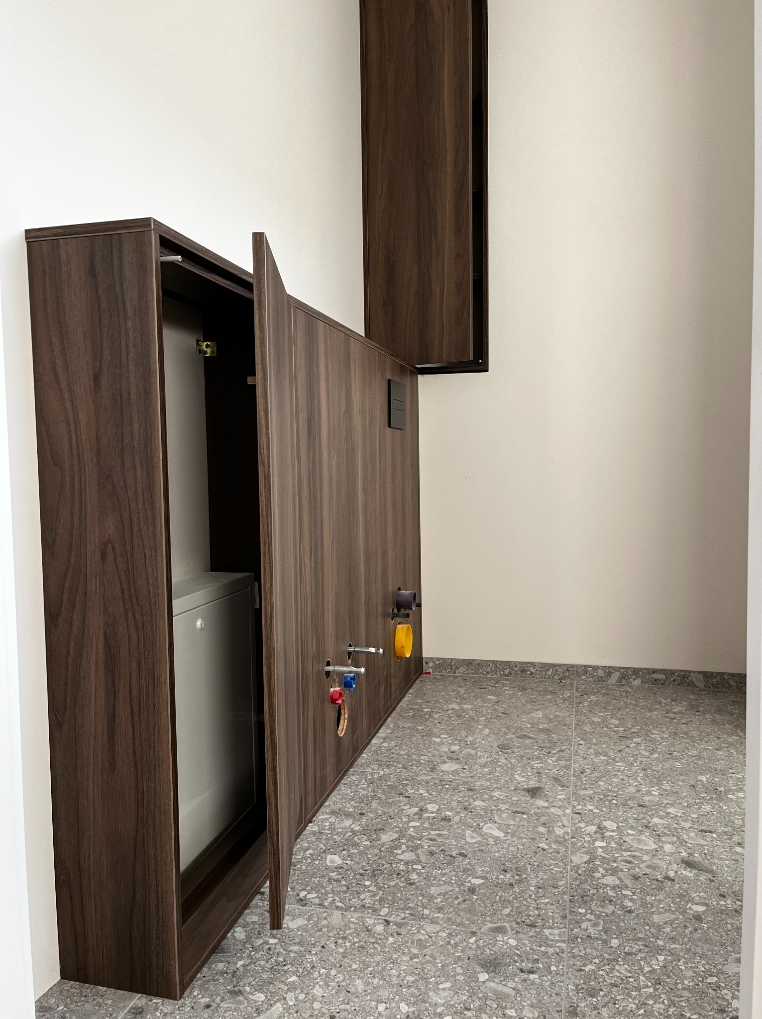 Szafa wnękowa z ciemnego drewna z otwartymi drzwiami, ukrywająca szary moduł. Widoczne elementy instalacji wodnej. Szara, lastrykowa podłoga.