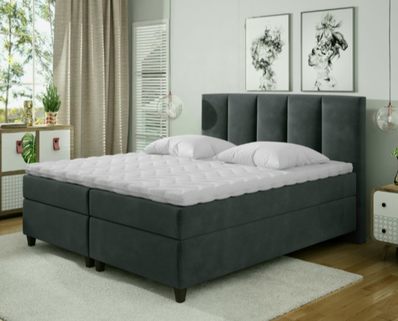 Szare łóżko tapicerowane z wysokim, pionowo pikowanym zagłówkiem, białymi poduszkami i materacem nawierzchniowym, na tle jasnego wnętrza z szafkami nocnymi i obrazami.