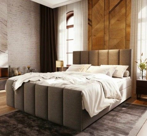 Tapicerowane łóżko kontynentalne w odcieniach szarości z wezgłowiem w pionowe pasy, pościelone białą pościelą, na tle ściany z drewnianą mozaiką i ceglanym murem.
