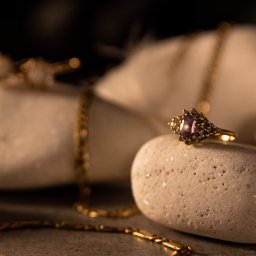 Jakub Kasperek - Złoty pierścionek z ametystem na porowatym kamieniu, w tle rozmyte złote łańcuszki i jasne kamienie. Ciepłe oświetlenie podkreśla detale biżuterii.