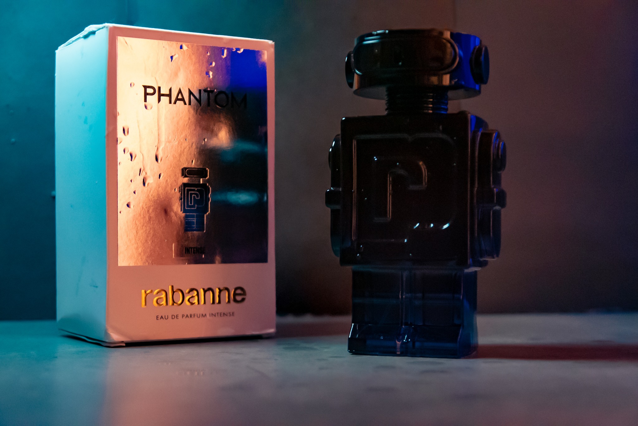 Flakon perfum Phantom Intense marki Rabanne w kształcie robota obok pudełka, oświetlony niebiesko-pomarańczowym światłem, na gładkiej powierzchni.