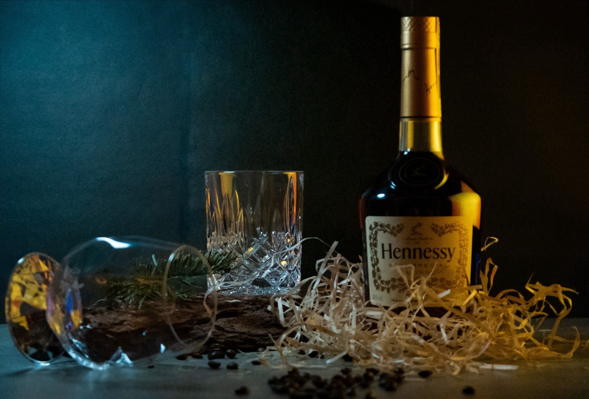 Butelka Hennessy obok szklanek z dekoracją, na ciemnym tle. Produkt w aranżacji z drewnem i kawą. Ciepłe, boczne oświetlenie podkreśla detale.