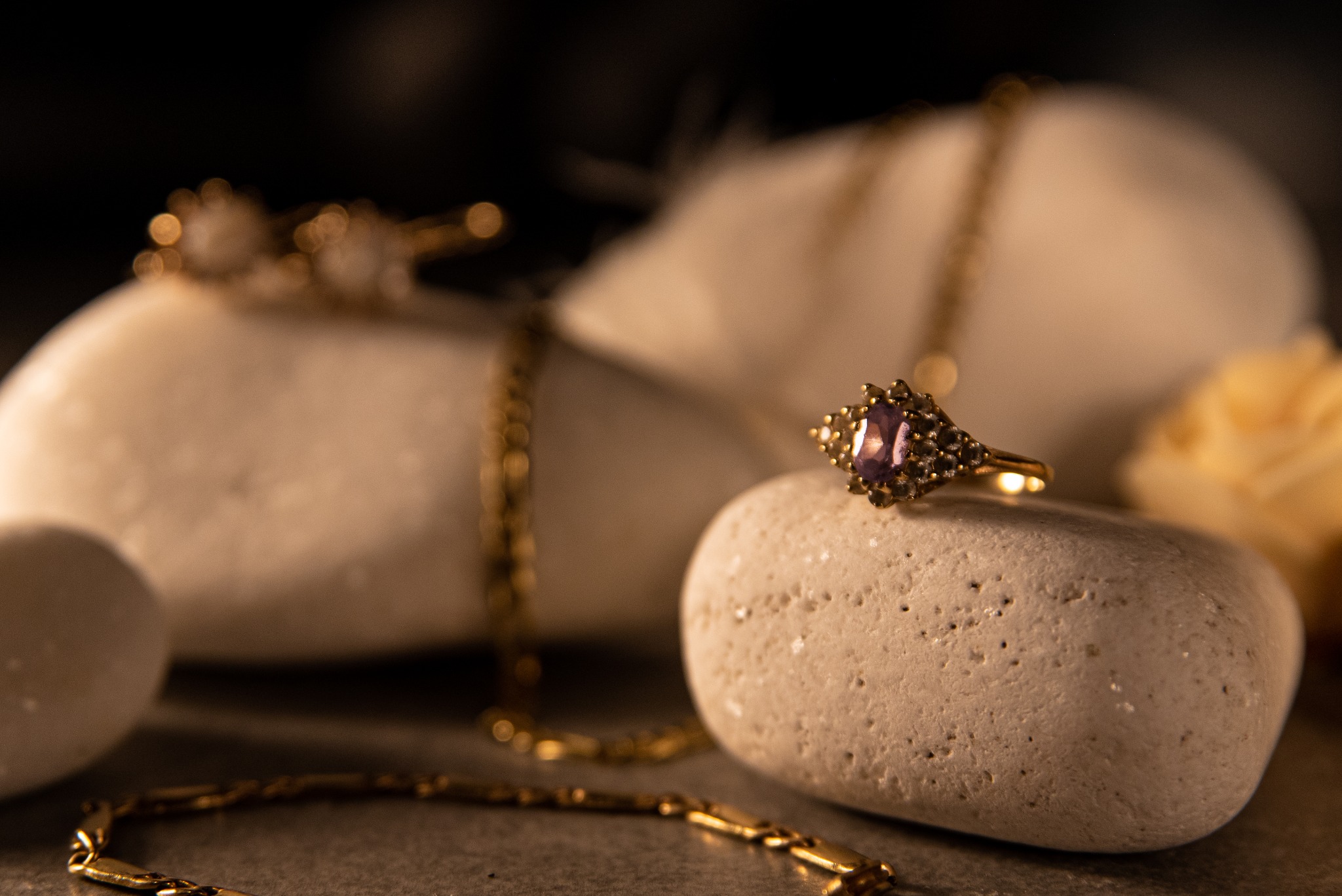 Złoty pierścionek z ametystem na porowatym kamieniu, w tle rozmyte złote łańcuszki i jasne kamienie. Ciepłe oświetlenie podkreśla detale biżuterii.