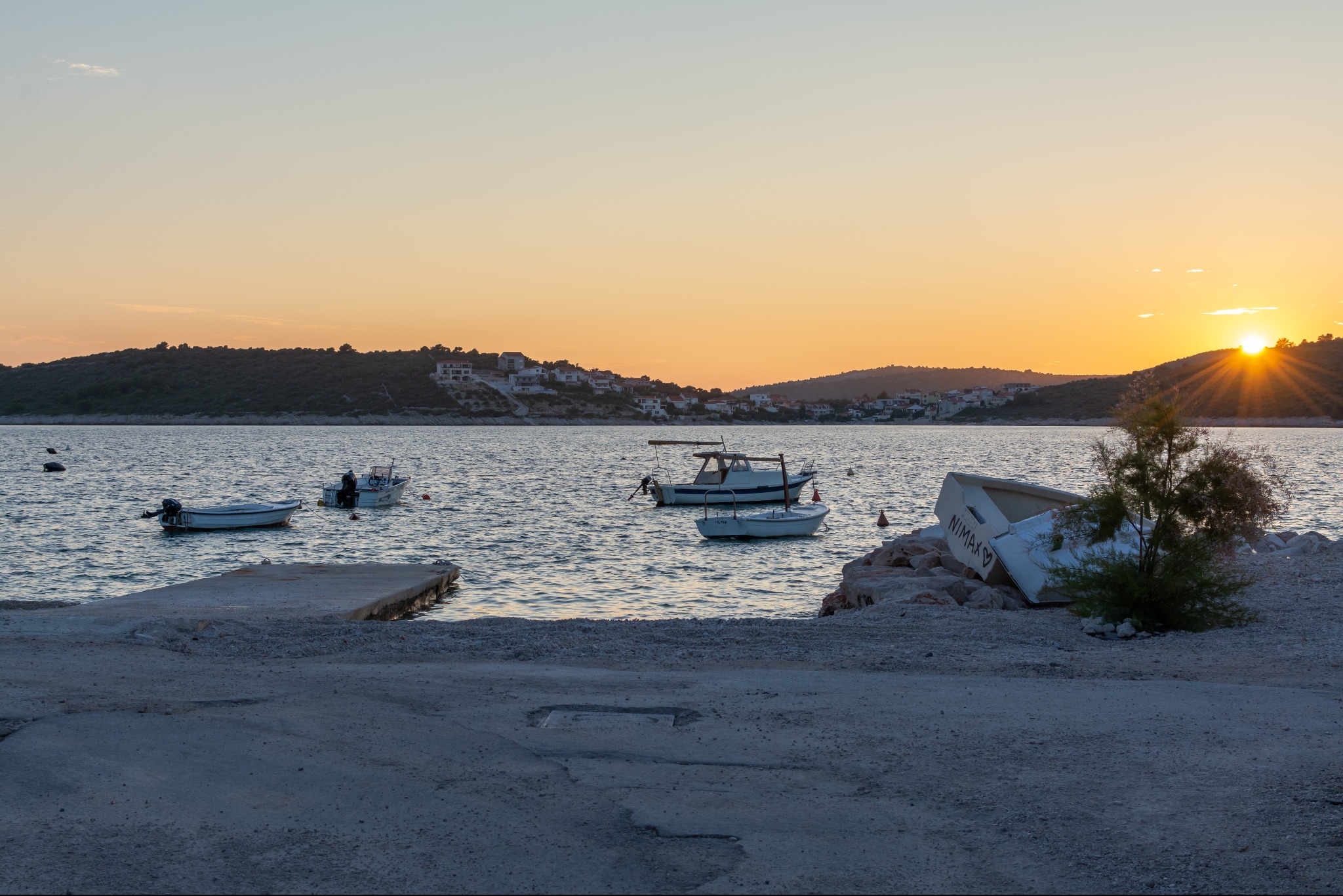 Zachód słońca nad morzem z łodziami, widok z betonowego nabrzeża. Przewrócona łódź na brzegu i budynki w tle. Ciepłe światło zachodzącego słońca.