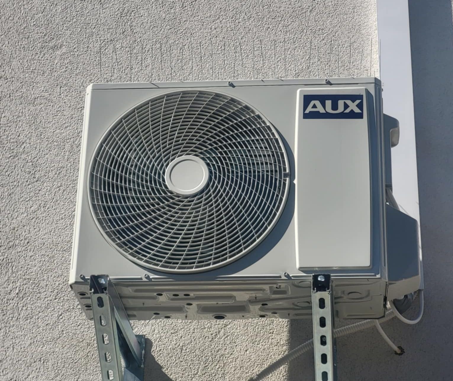 Zewnętrzna jednostka klimatyzacji AUX zamontowana na szarej, teksturowanej ścianie budynku. Widoczne metalowe wsporniki i przewody. Ujęcie z dołu.