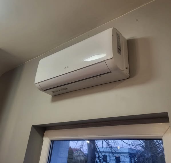 Biały klimatyzator AUX zamontowany na szarej ścianie nad oknem. Widoczny fragment okna z widokiem na zewnątrz. Neutralne oświetlenie, minimalistyczny design.