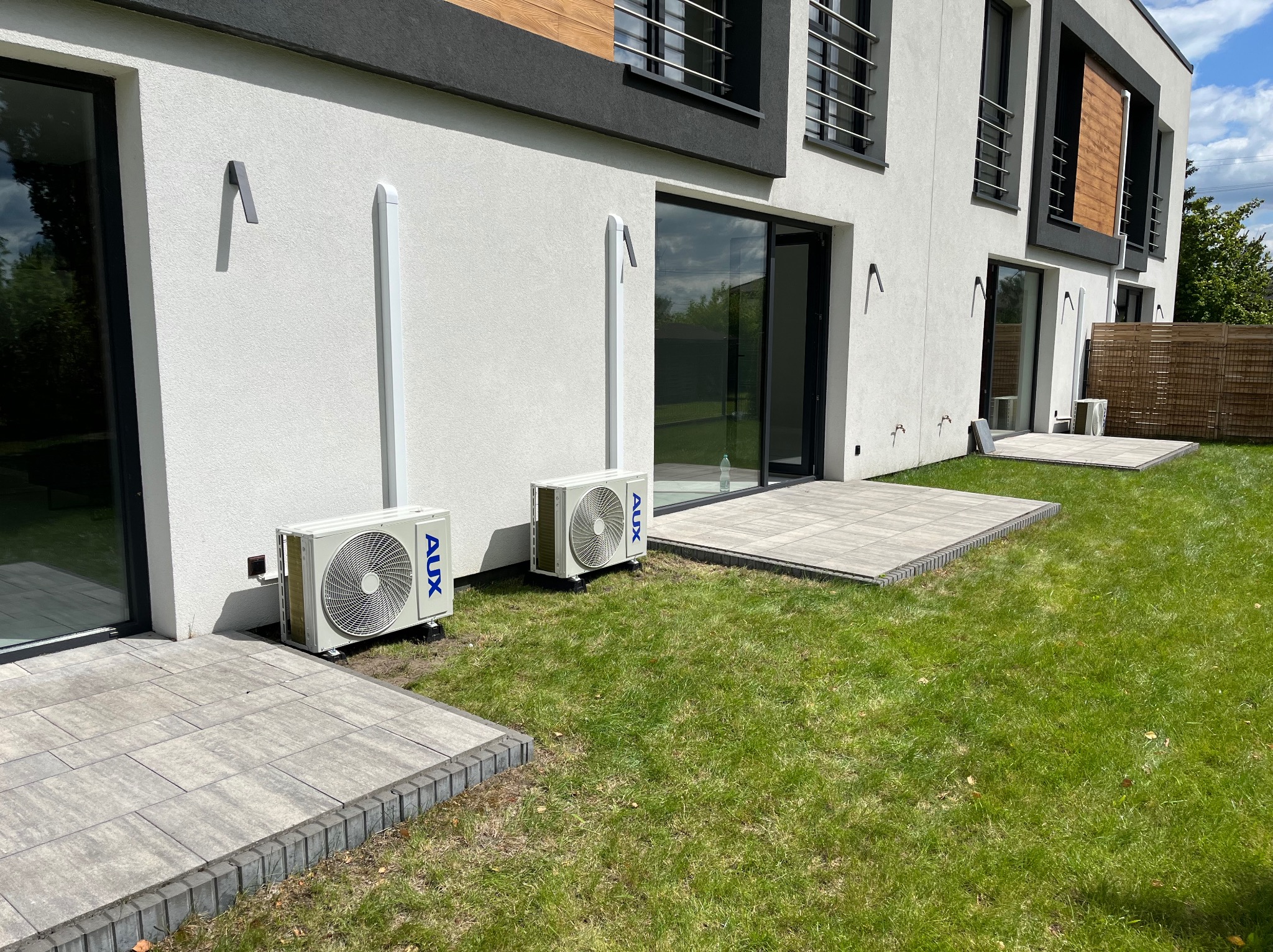 Dwa białe klimatyzatory AUX zamontowane na zewnątrz nowoczesnego domu z szarymi elementami elewacji i dużymi oknami, na tle zielonego trawnika i kostki brukowej.