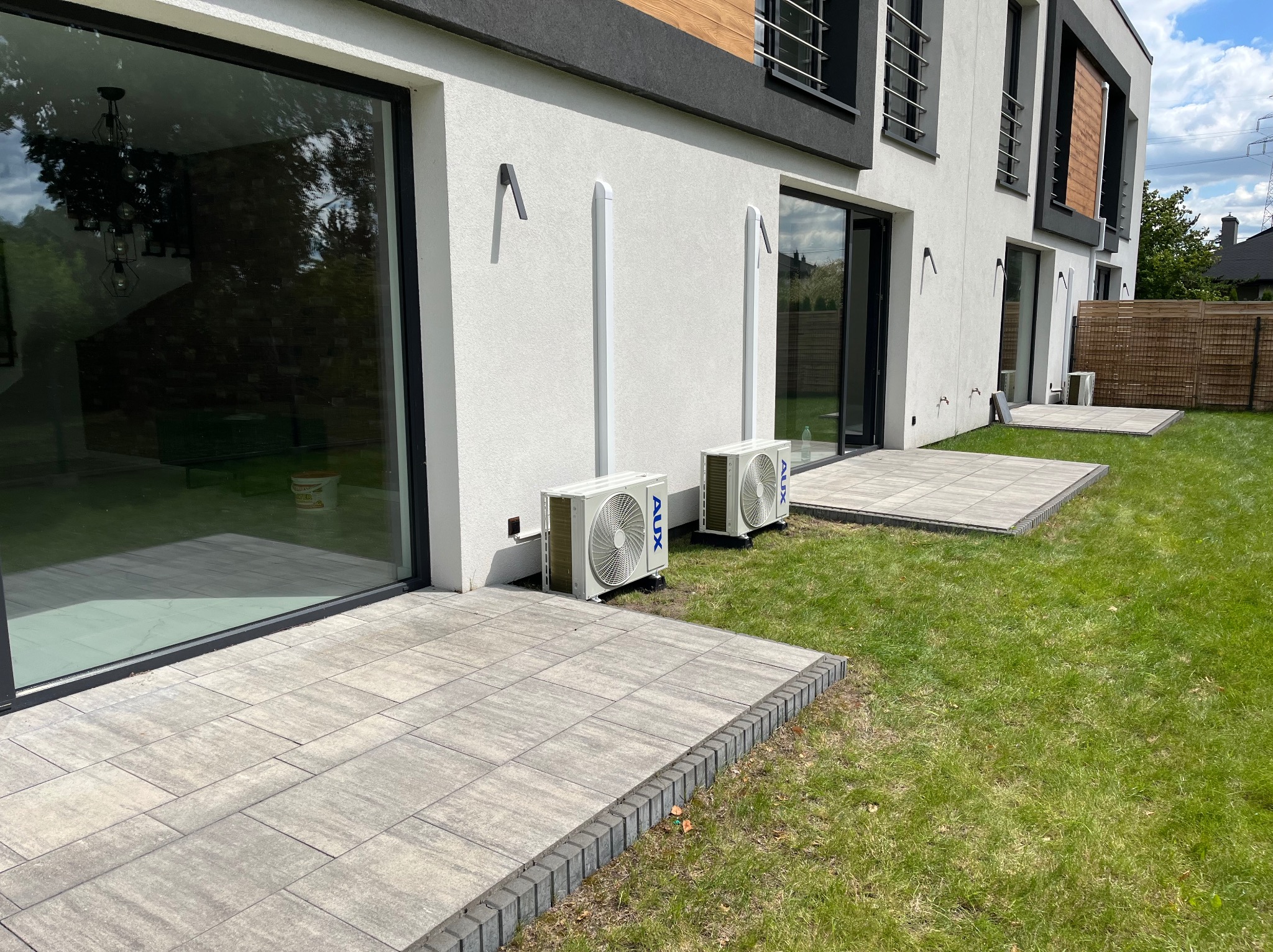 Dwa klimatyzatory AUX zamontowane na elewacji nowoczesnego domu z wyjściem na taras wyłożony kostką. Widoczne rury odprowadzające skropliny.