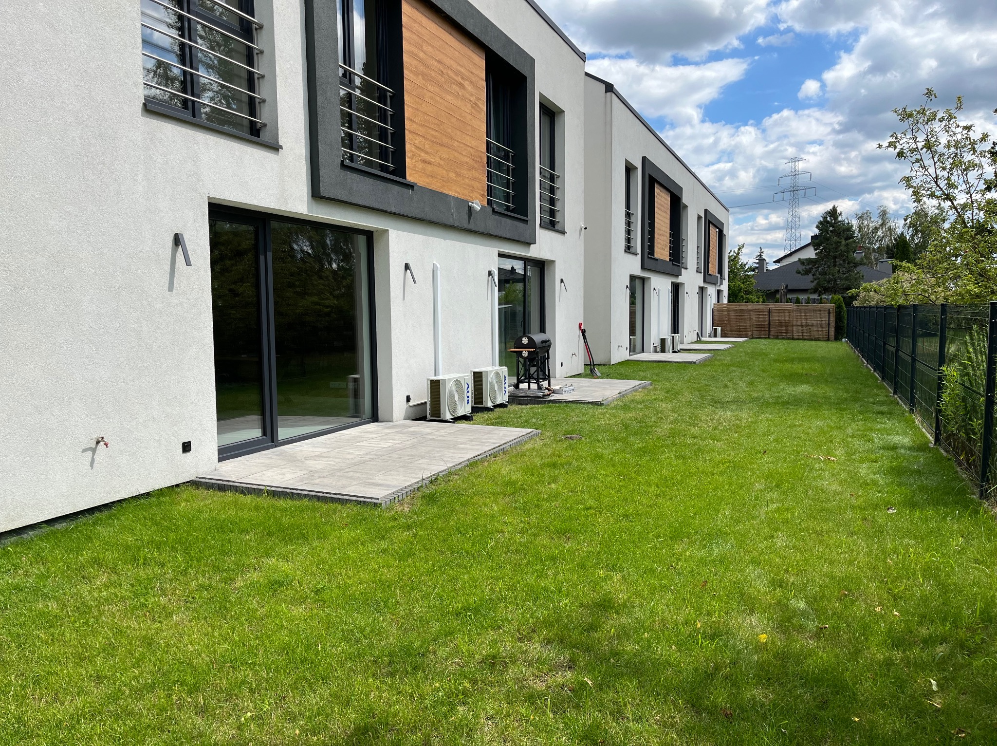 Biały budynek szeregowy z zamontowanymi klimatyzatorami na zewnątrz, widok z zadbanego trawnika w słoneczny dzień.