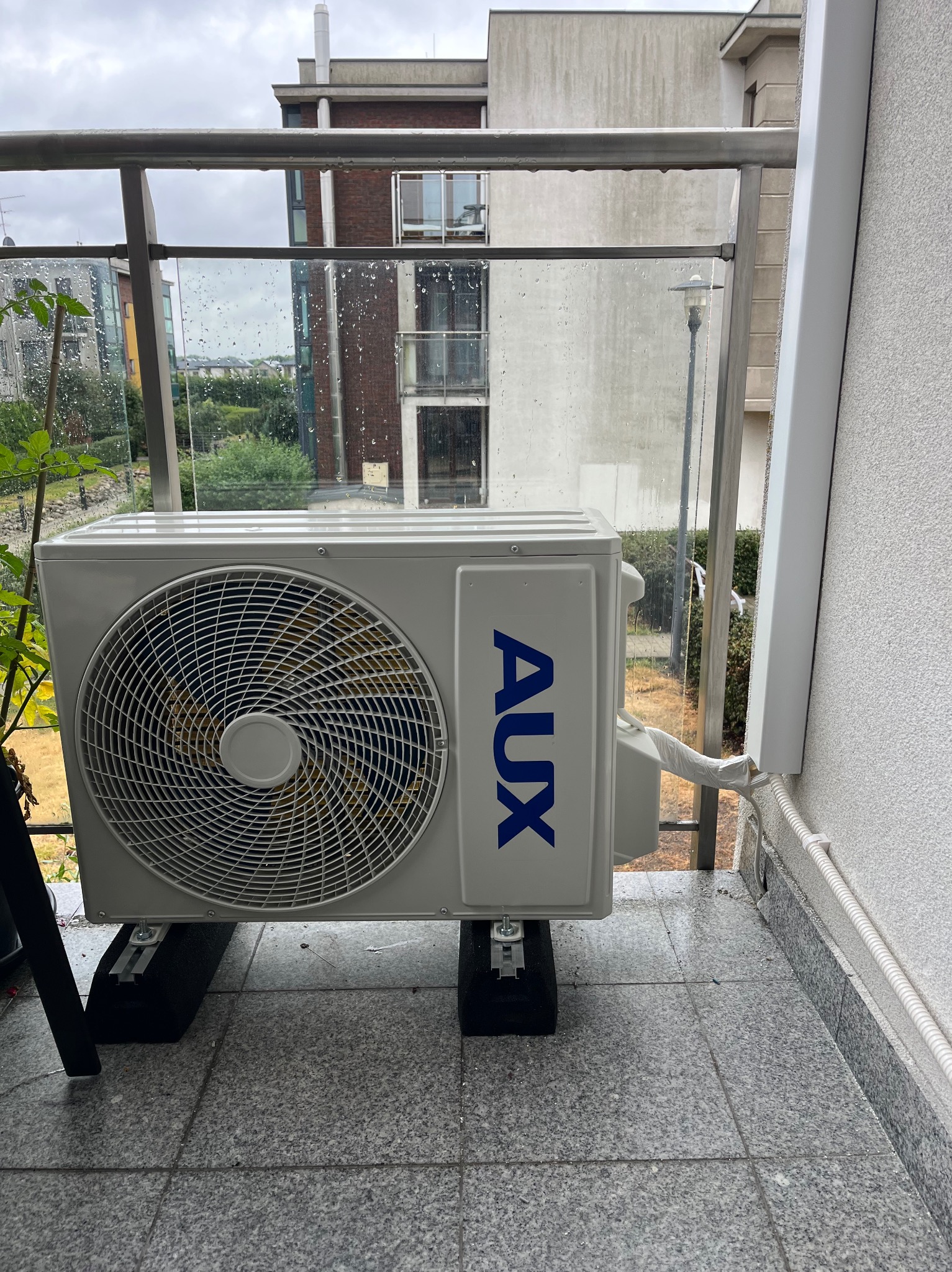 Klimatyzator AUX zamontowany na balkonie w Piasecznie, widok z bliska. Urządzenie stoi na czarnych podkładkach, tło stanowią budynki w deszczu za szklaną balustradą.