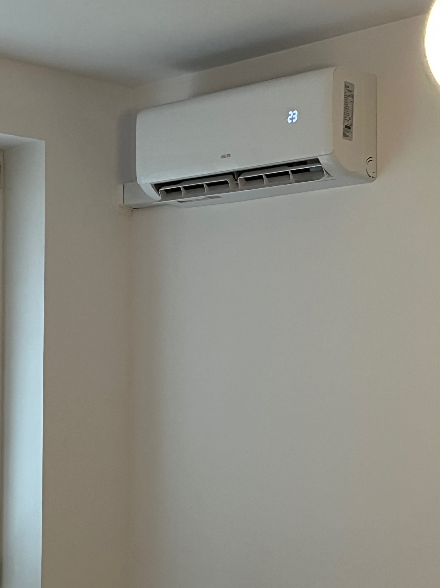 Biała klimatyzacja AUX zamontowana na ścianie w jasnym pomieszczeniu, wyświetlająca temperaturę 23 stopnie. Widoczny fragment okna po lewej stronie.