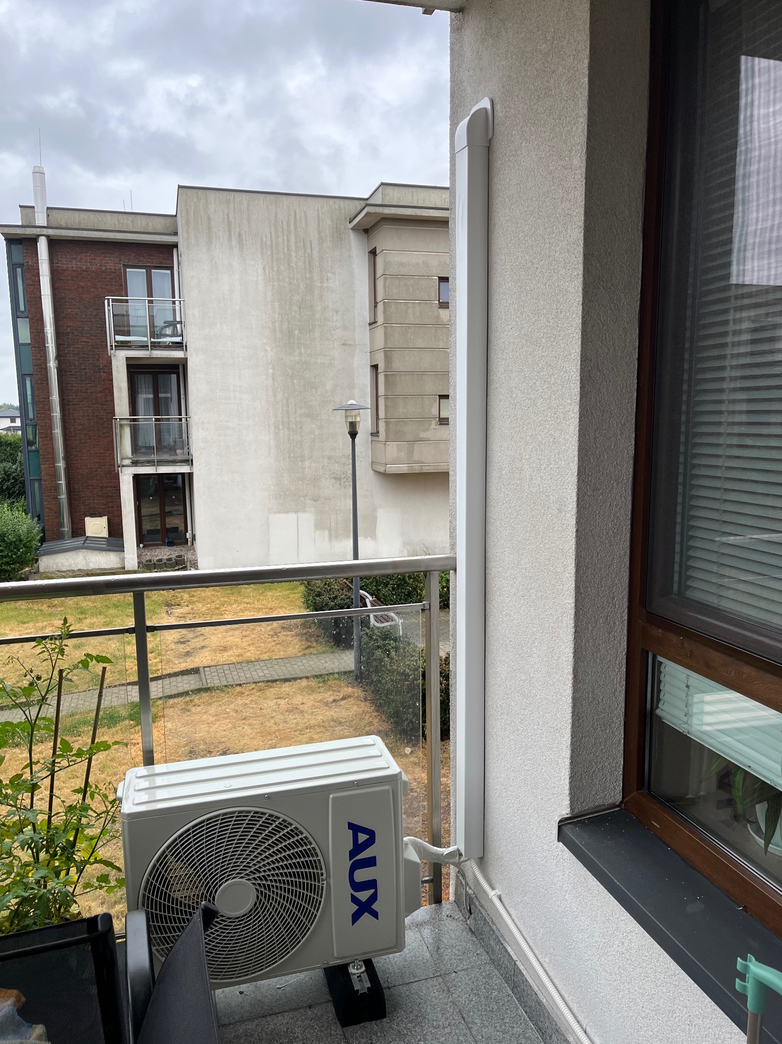 Biała jednostka zewnętrzna klimatyzacji AUX na balkonie z szarą podłogą, widoczna rura odprowadzająca na elewacji budynku. Ujęcie z bliska, w tle zabudowa miejska.