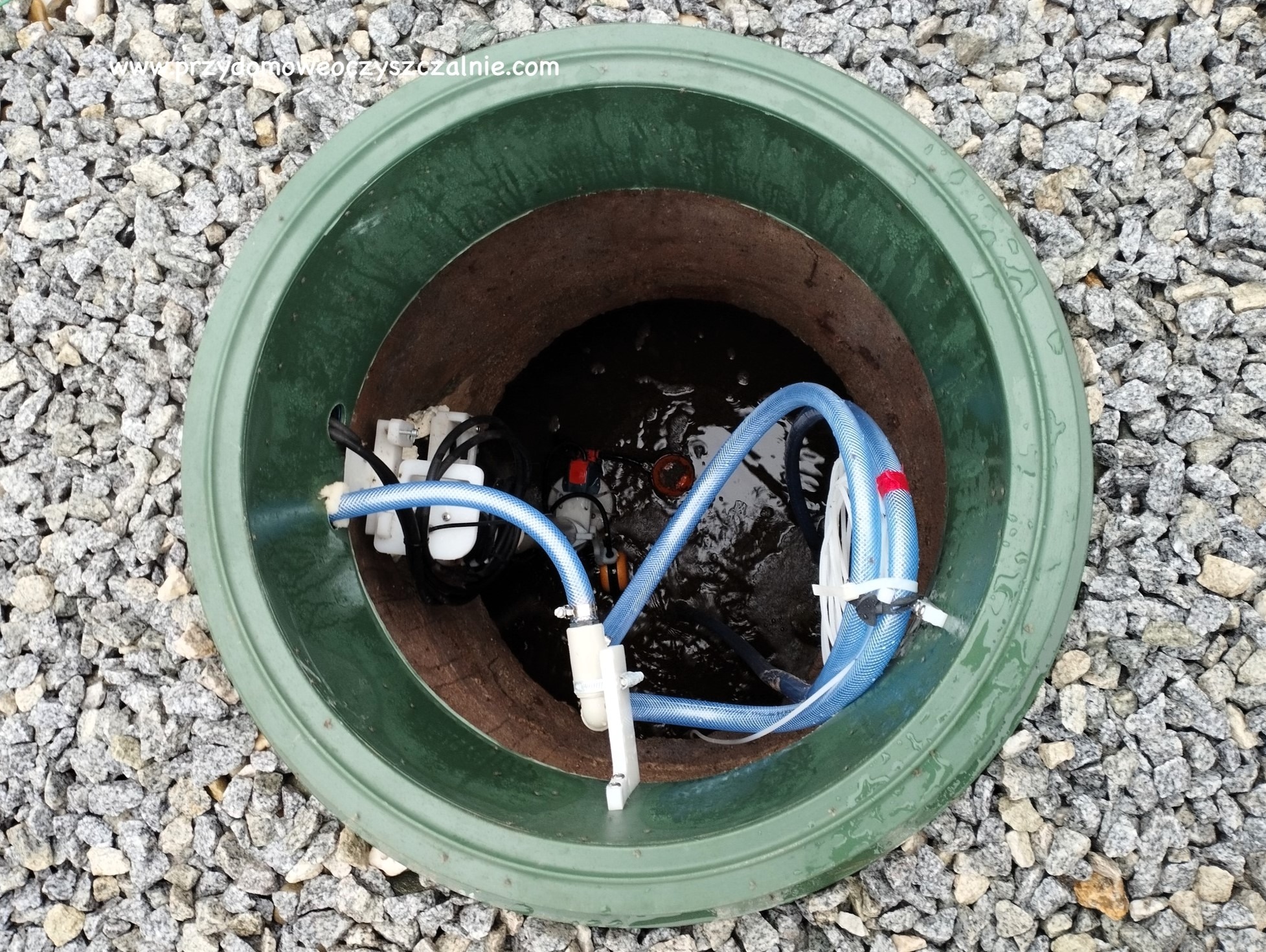Zielona studzienka rewizyjna przydomowej oczyszczalni ścieków z widocznymi elementami instalacji wewnętrznej, takimi jak pompy i węże, otoczona drobnym, szarym żwirem.