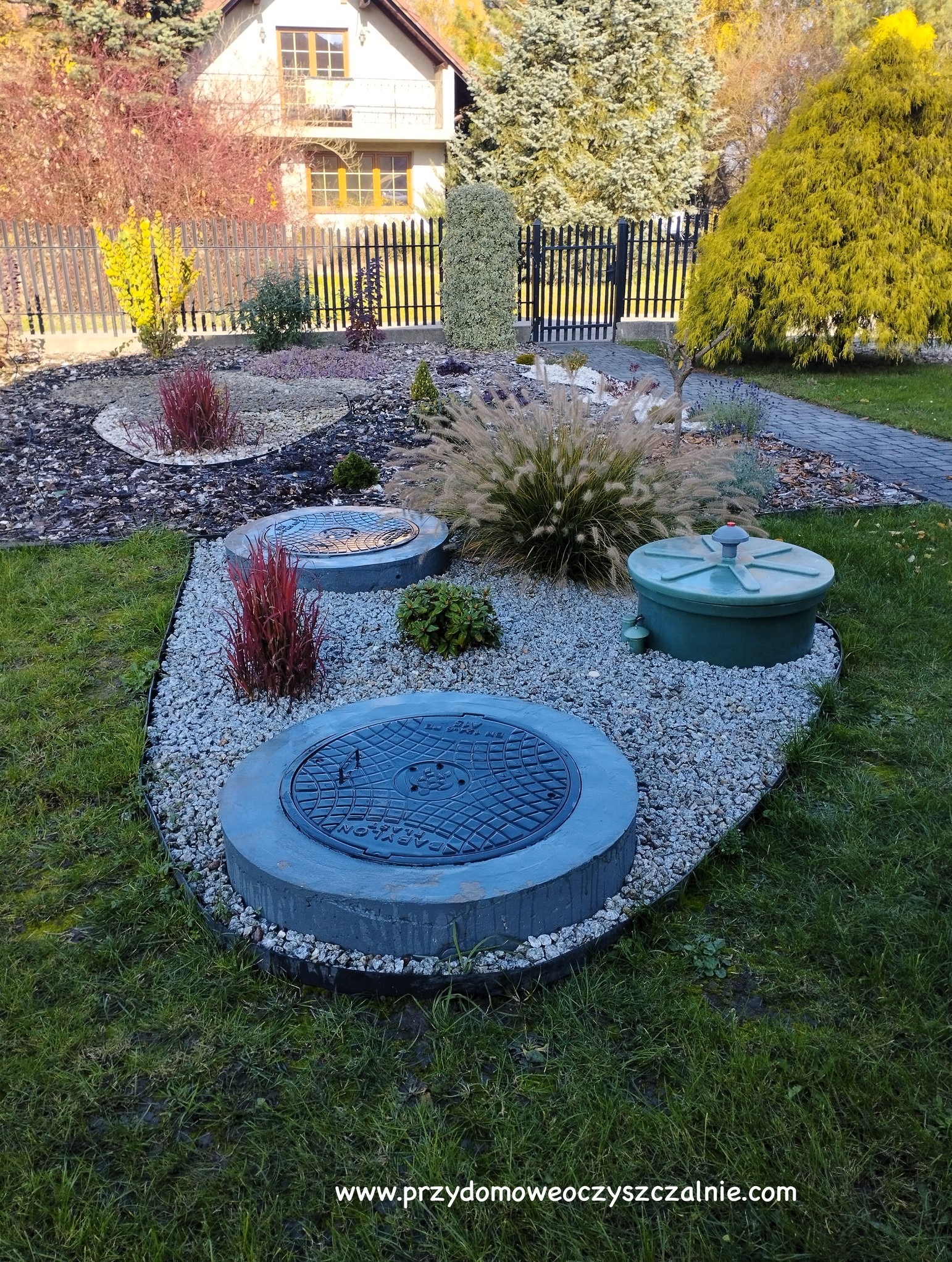 Trzy betonowe studzienki rewizyjne z żeliwnymi pokrywami i zielony zbiornik na tle ogrodu z ozdobnymi trawami i kamieniami, widok z góry.