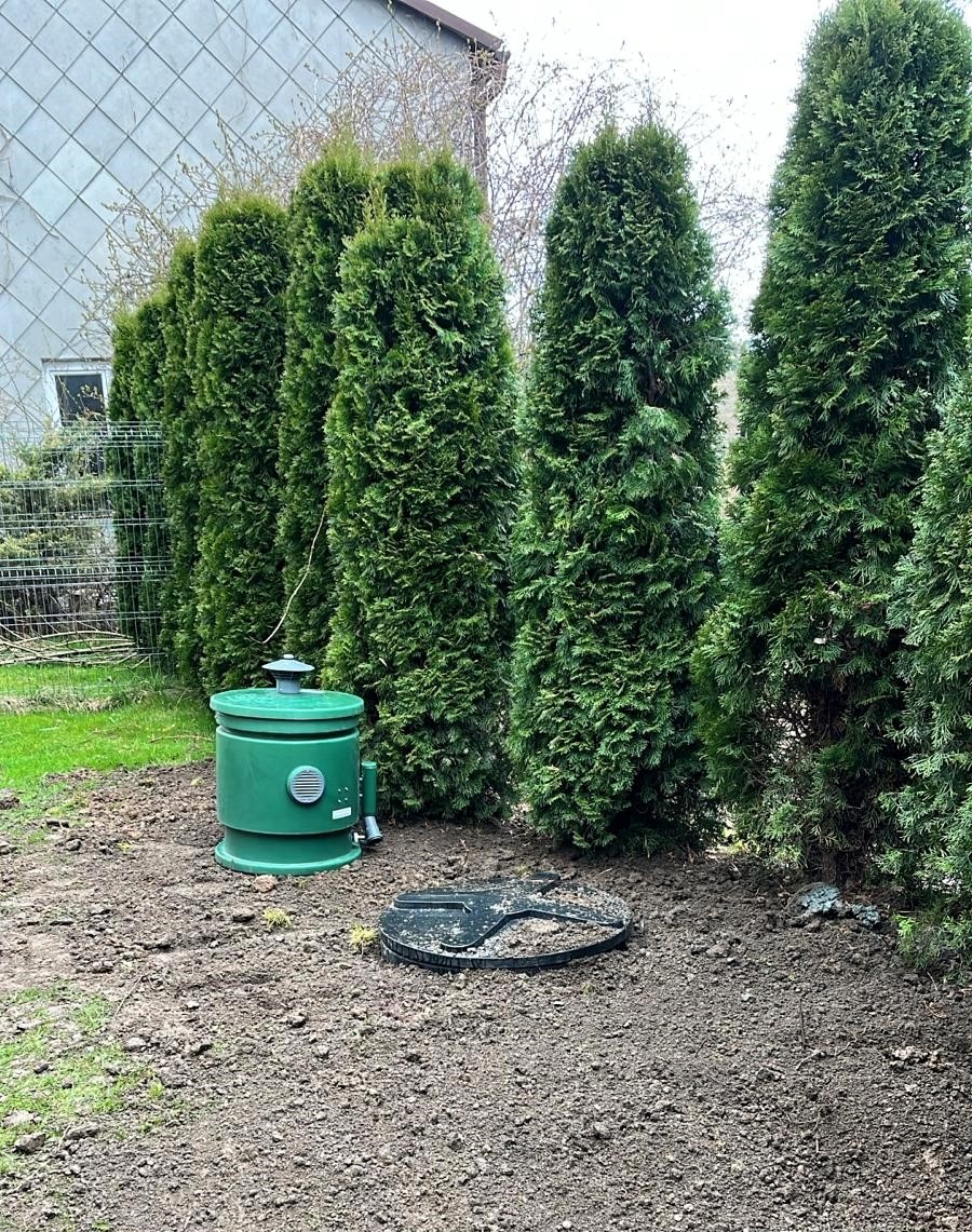 Zielona przydomowa oczyszczalnia ścieków z odpowietrznikiem i pokrywą włazu na tle rzędu tuj w ogrodzie.