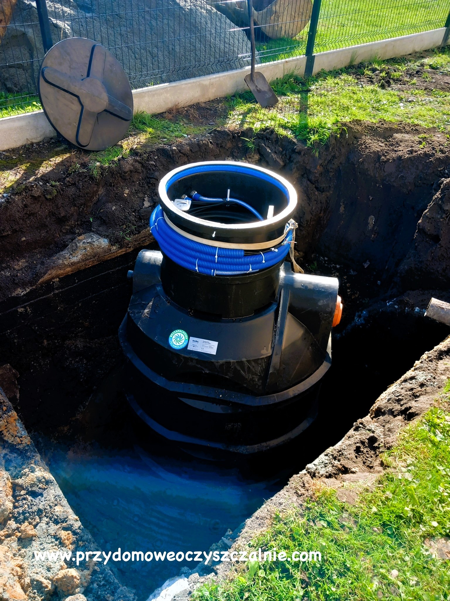 Montaż czarnej, plastikowej oczyszczalni ścieków w wykopie w Tychach, widoczna niebieska rura karbowana i pokrywa obok wykopu. Szpadel oparty o ogrodzenie w tle.