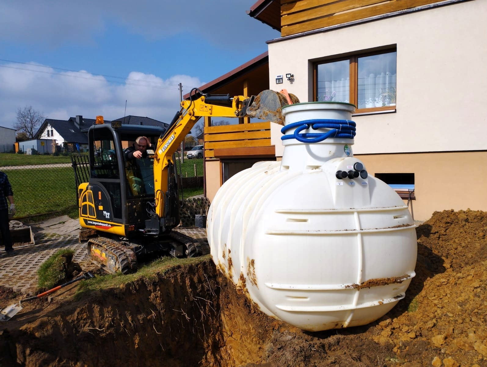 Żółta mini koparka umieszcza biały zbiornik z niebieskimi rurami w wykopie obok domu jednorodzinnego.