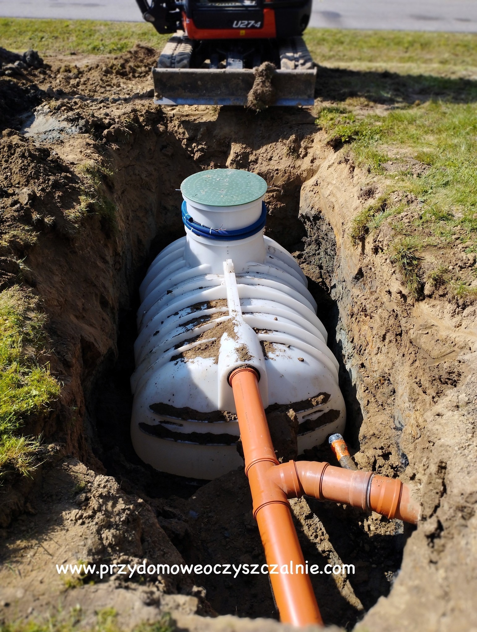 Biały zbiornik z zieloną pokrywą i niebieskim wężem karbowanym, umieszczony w wykopie ziemnym z widocznymi rurami kanalizacyjnymi z pomarańczowego tworzywa, na tle minikoparki.