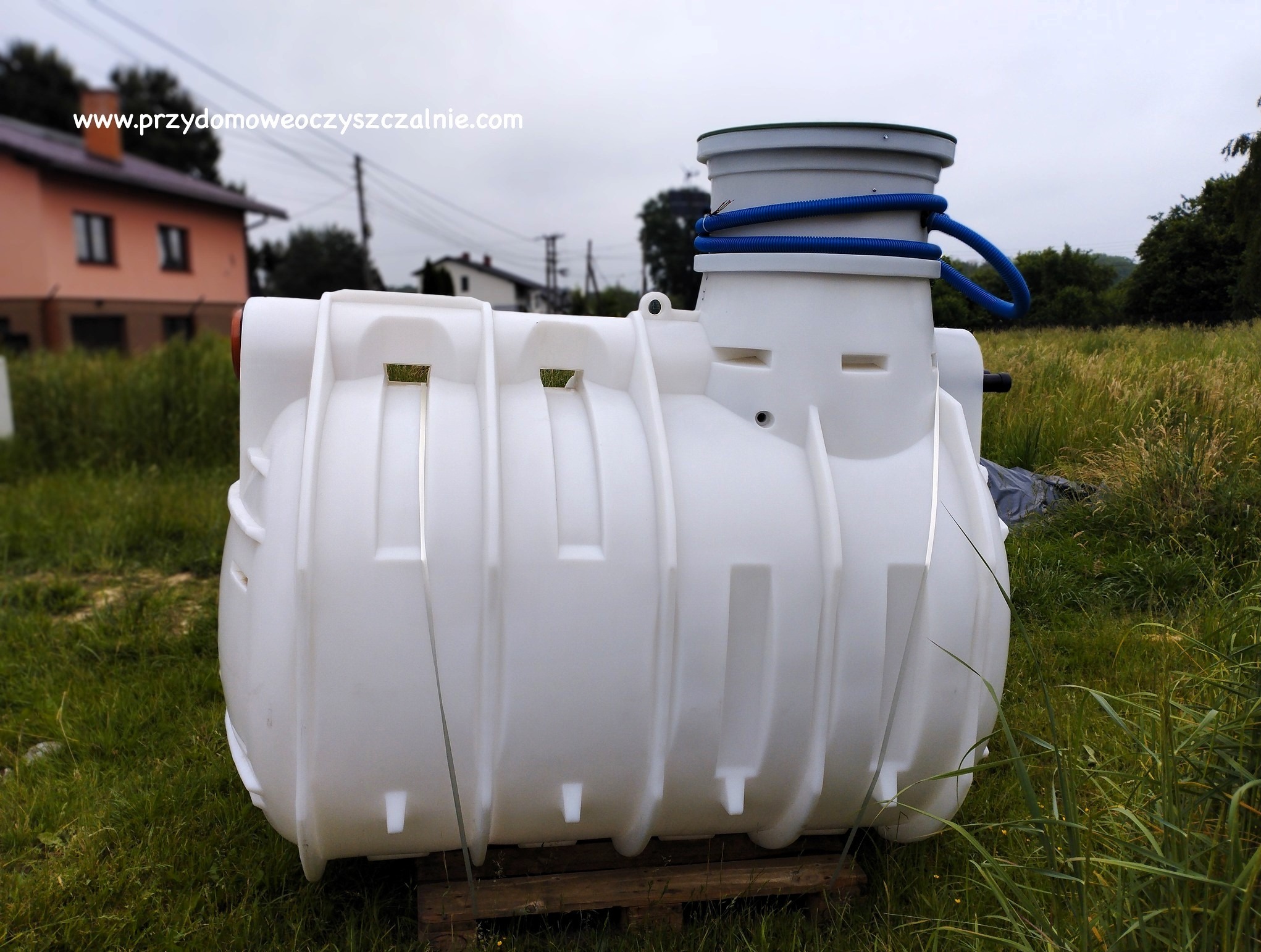 Biały zbiornik przydomowej oczyszczalni ścieków z niebieskim przewodem, umieszczony na drewnianej palecie na tle trawy i zabudowań.