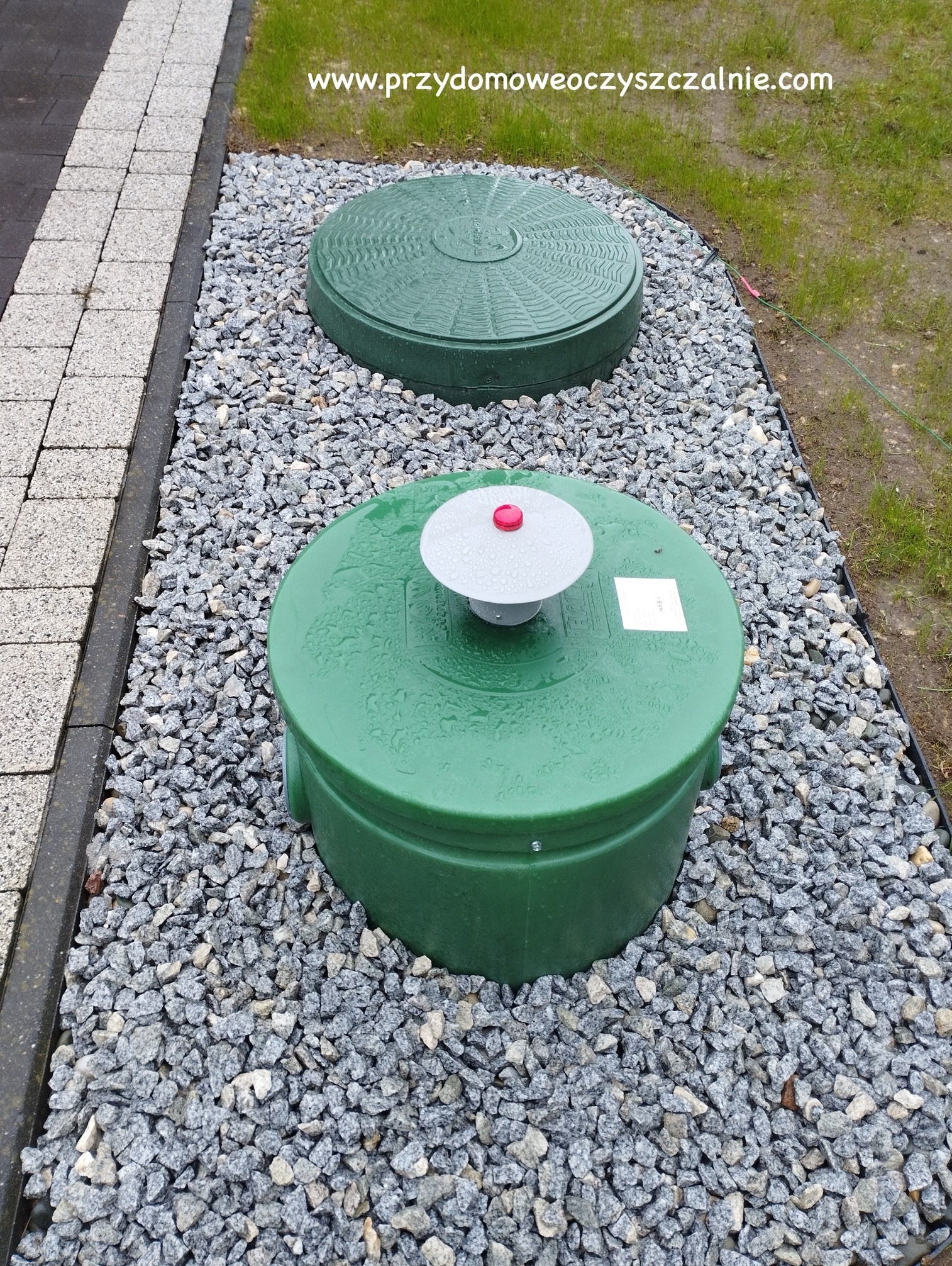 Dwa zielone włazy inspekcyjne, jeden z odpowietrznikiem z czerwoną nakładką, otoczone szarym grysem, na tle trawnika i kostki brukowej.