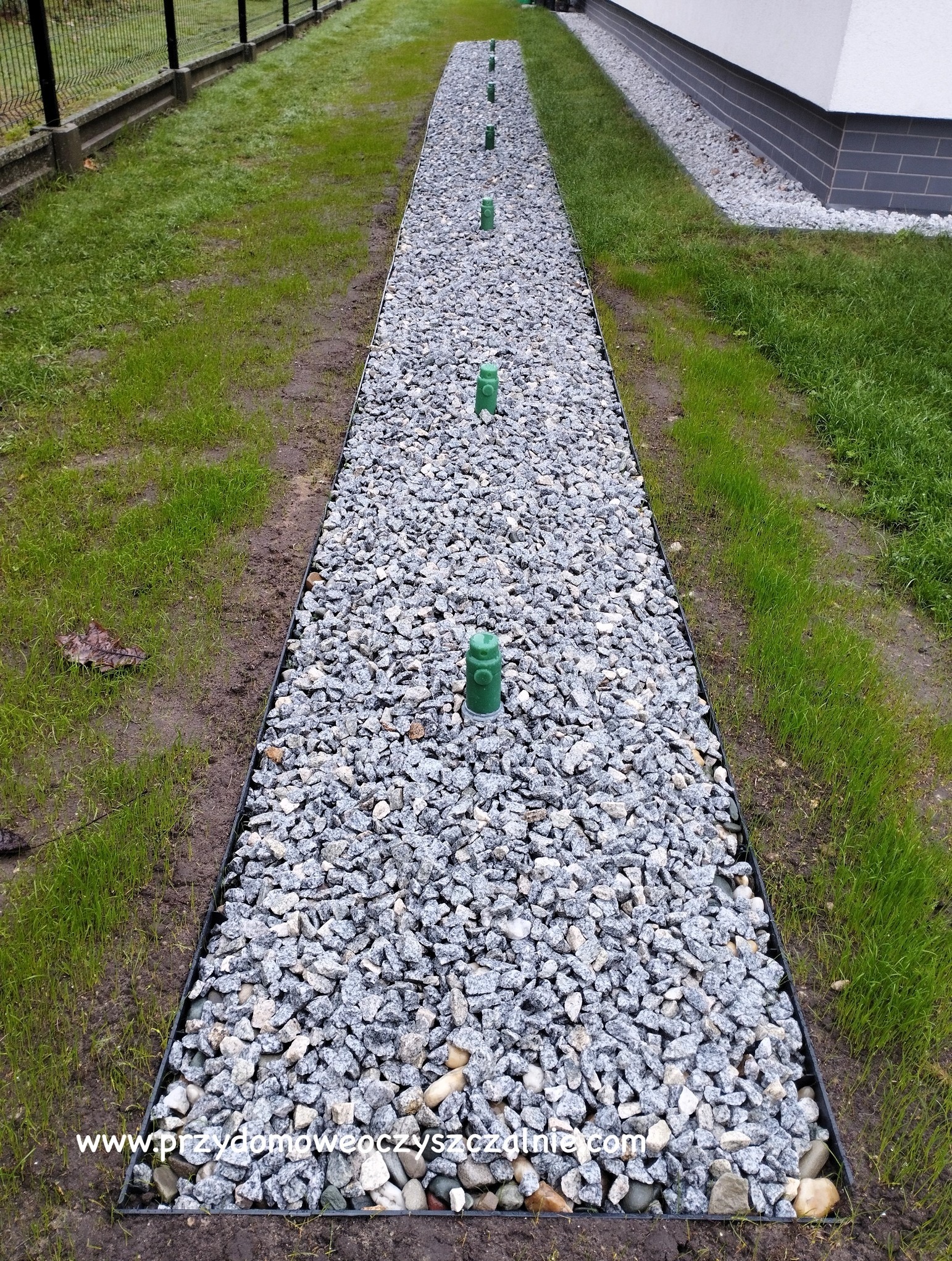 Długi drenaż rozsączający obsypany szarym grysem, z pionowymi zielonymi odpowietrznikami, obrzeżony czarną listwą, widok z góry wzdłuż trawnika i budynku.