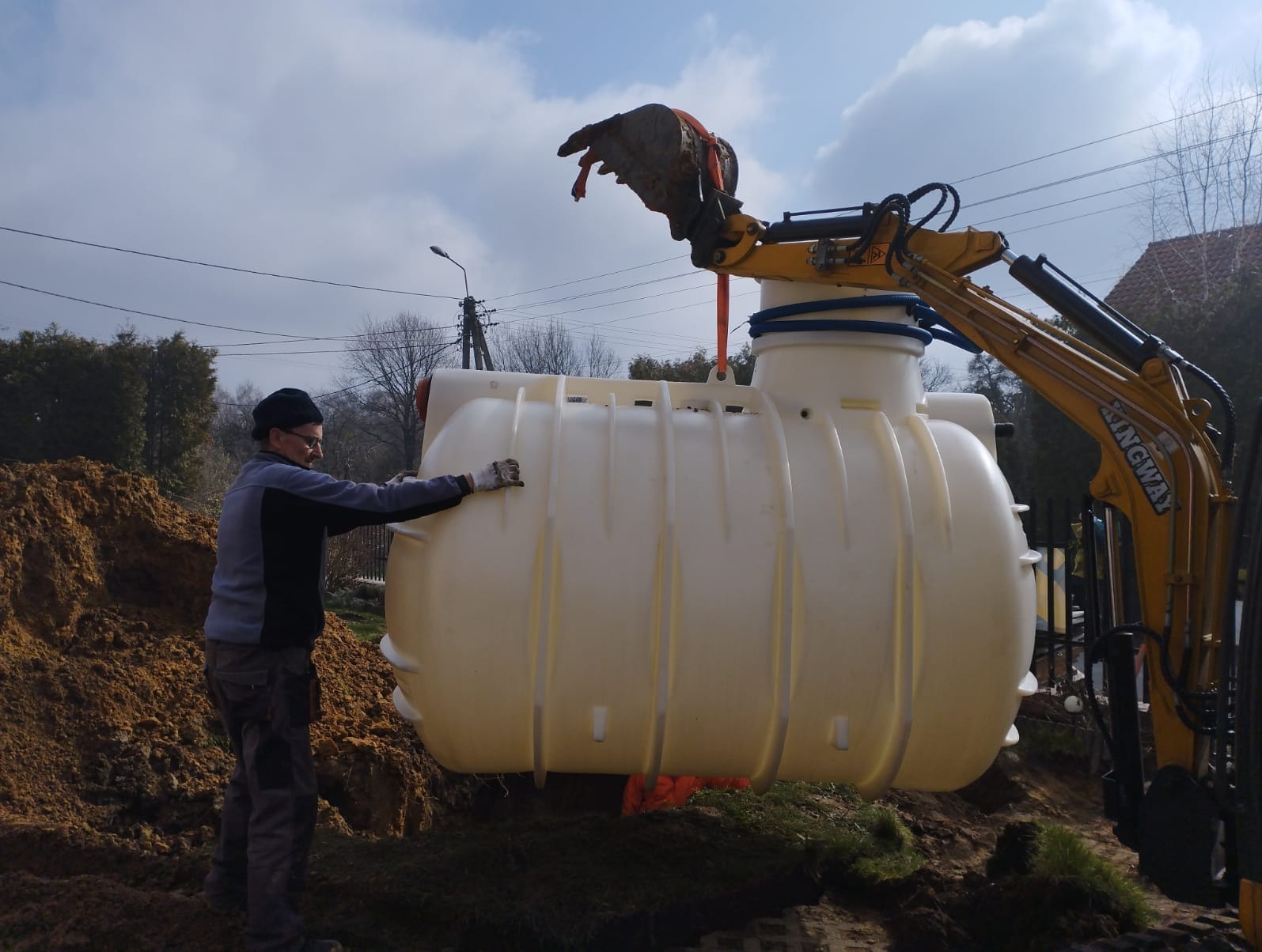 Montaż beżowej, poziomej oczyszczalni ścieków z pomocą żółtej mini-koparki KINGWAY; pracownik w rękawicach stabilizuje zbiornik przed opuszczeniem do wykopu.