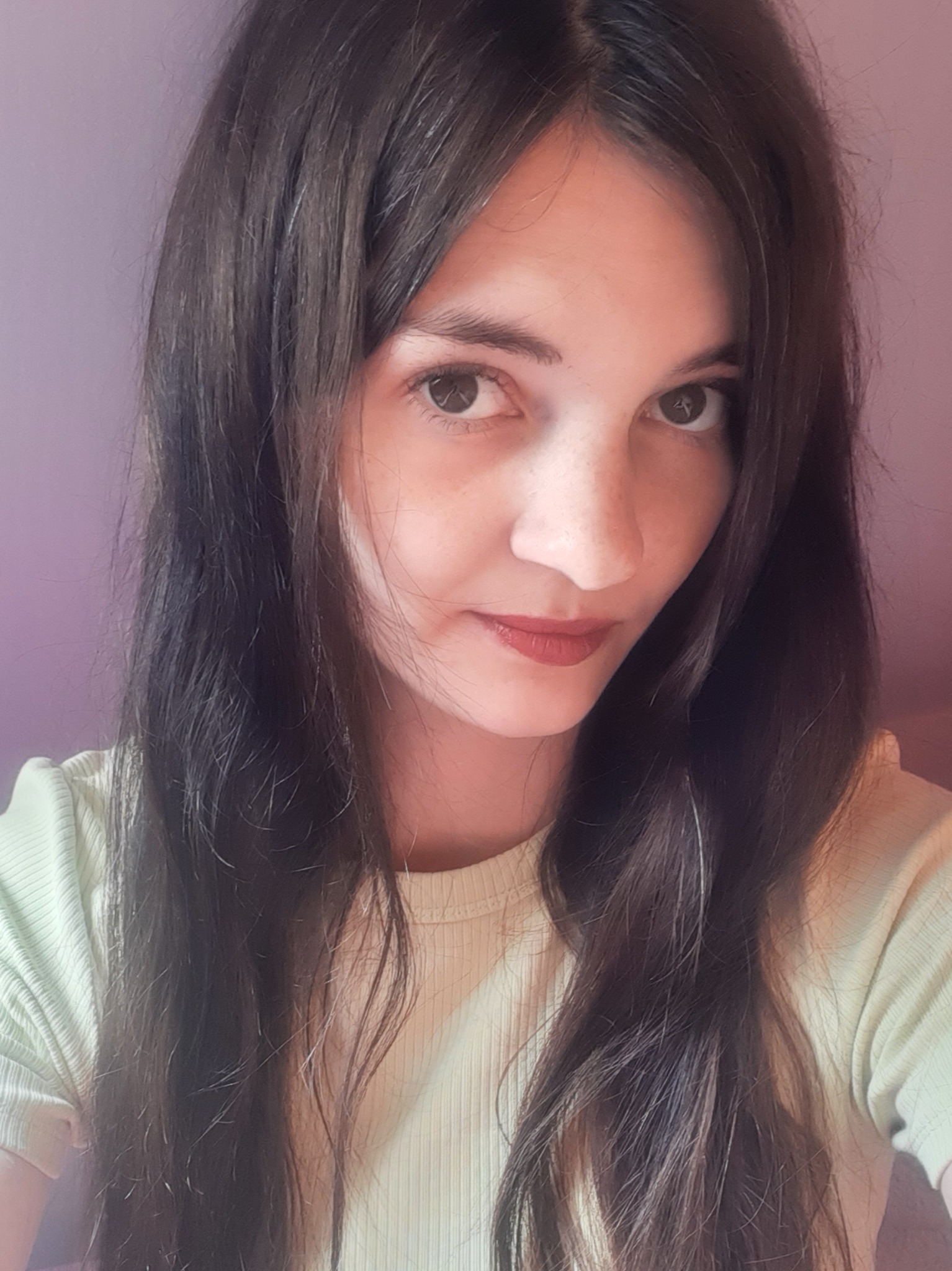 Portret kobiety o brązowych włosach i oczach, z delikatnym uśmiechem na tle jasnofioletowej ściany, ubrana w jasnozieloną koszulkę.