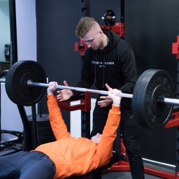 KTP KATOWICE SPÓŁKA Z OGRANICZONĄ ODPOWIEDZIALNOŚCIĄ - Mężczyzna w pomarańczowej bluzie wyciska sztangę na ławeczce pod okiem instruktora w czarnej bluzie z kapturem w siłowni.