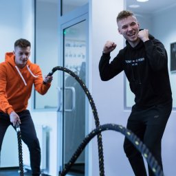 KTP KATOWICE SPÓŁKA Z OGRANICZONĄ ODPOWIEDZIALNOŚCIĄ - Dwóch mężczyzn ćwiczy na siłowni z linami treningowymi, jeden w pomarańczowej bluzie wykonuje ćwiczenie, drugi w czarnej bluzie z uniesionymi pięściami w geście radości, w tle worek treningowy...
