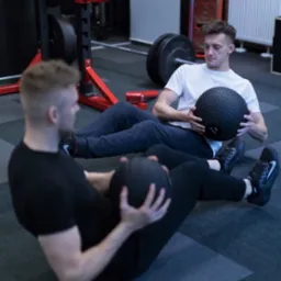 Dwóch mężczyzn wykonuje ćwiczenia z piłkami lekarskimi w siłowni, z widocznym sprzętem do podnoszenia ciężarów w tle.