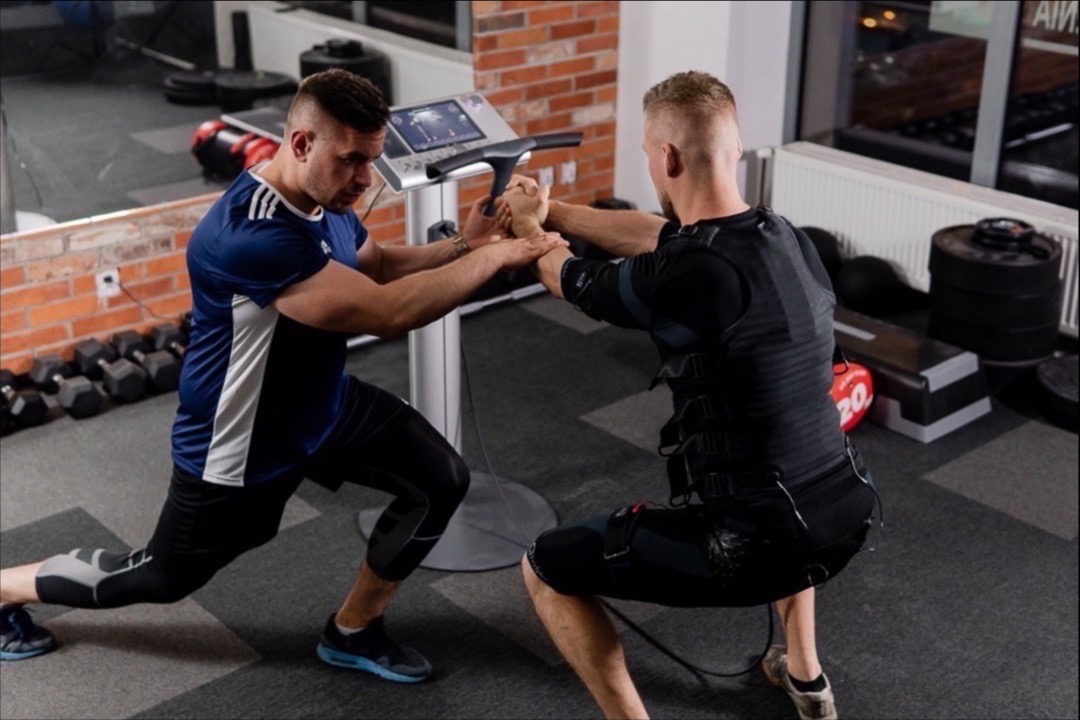 Trening EMS: mężczyzna w stroju do elektrostymulacji mięśni wykonuje przysiad z pomocą trenera, który stabilizuje jego pozycję przy maszynie z wyświetlaczem.